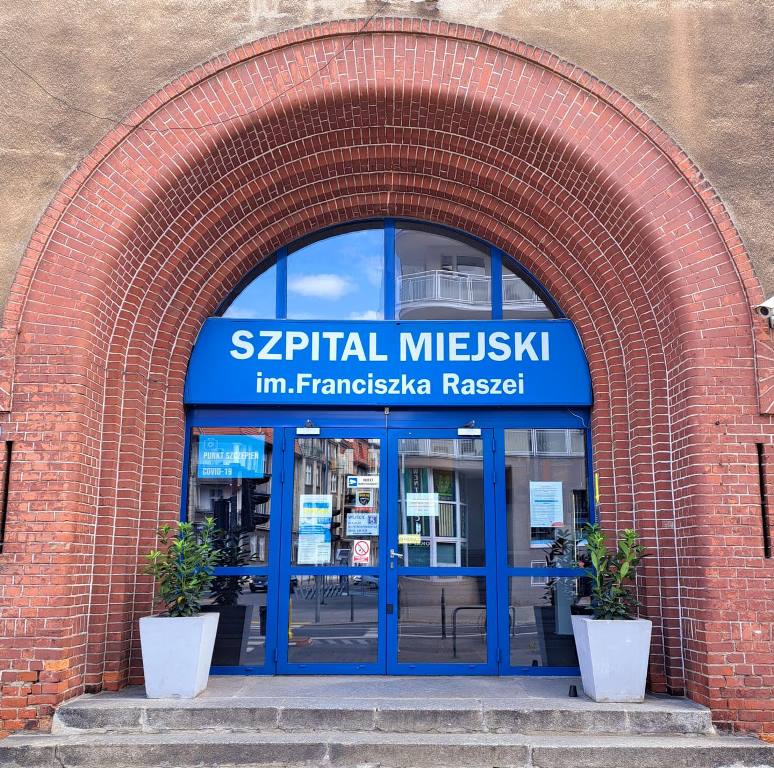 wejście do budynku w formie ceglanego łuku drzwi wejściowe są przeszklone z niebieskimi framigami nad drzwiami napsi SZPITAL MIEJSKI im. Franciszka Raszei przed wejściem stoją dwie donice z roślinami