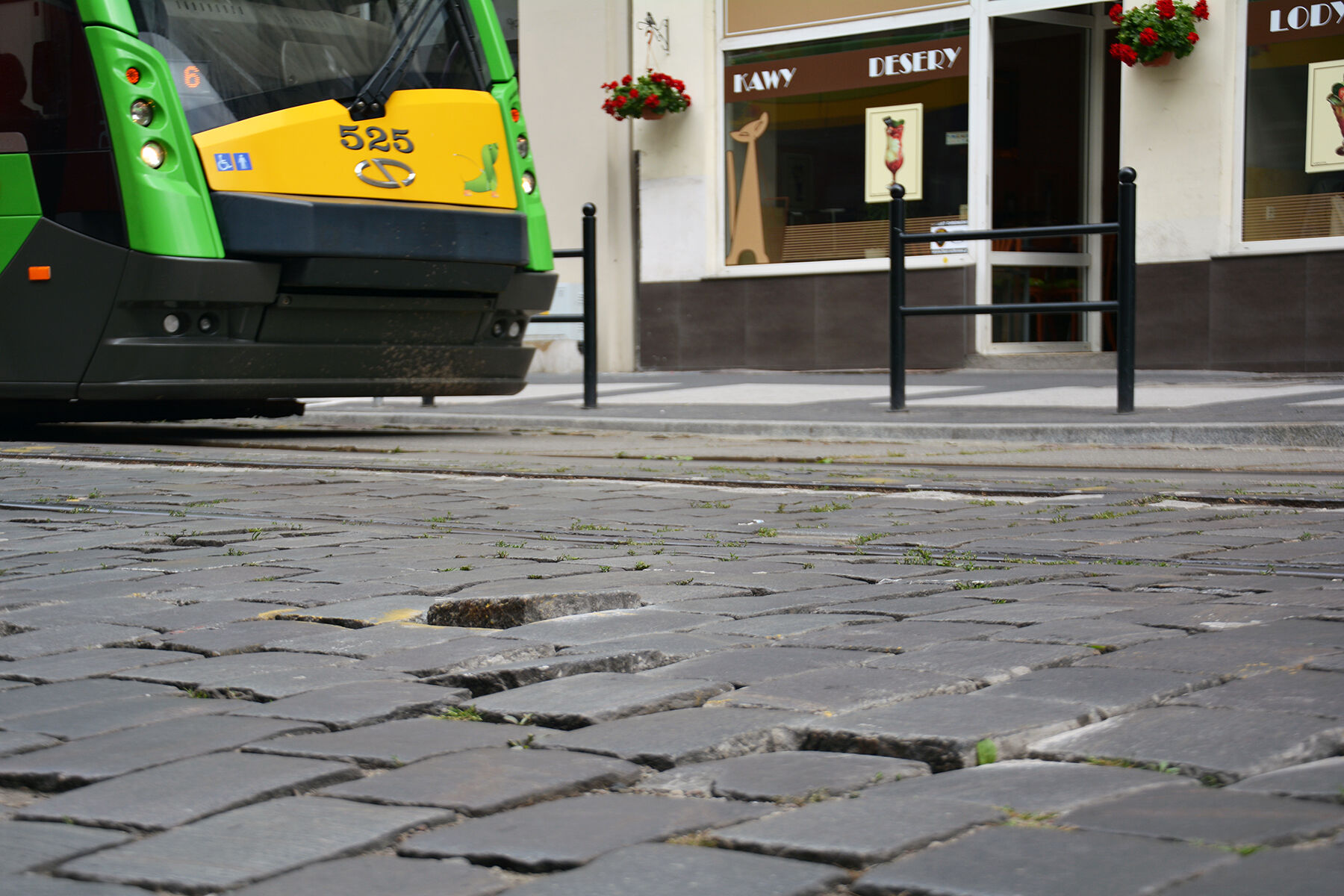 Widać nawierzchnię z nierówną kostką, w tle część tramwaju i sklepy.