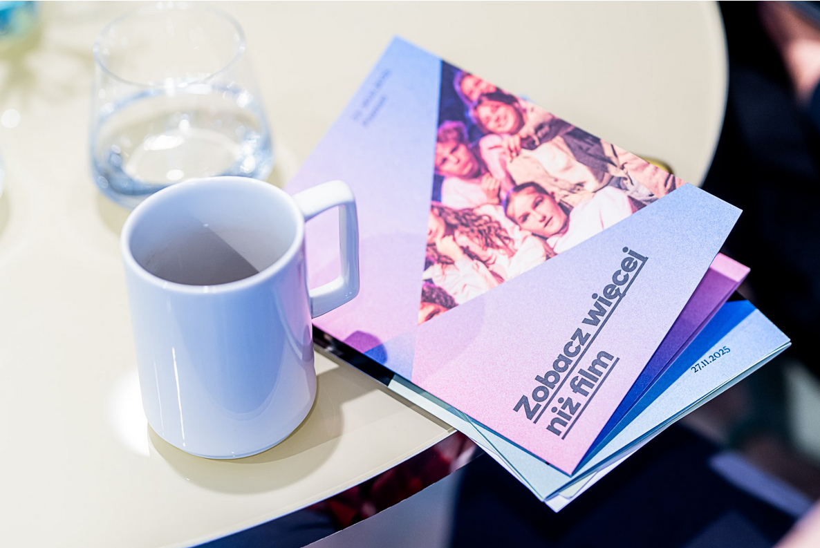 Widoczny od góry stół, na nim kubek, szklanka z wodą oraz wydrukowany program festiwalu z napisem: "zobacz więcej niż film" i datą wydarzenia, zawartą w treści artykułu.