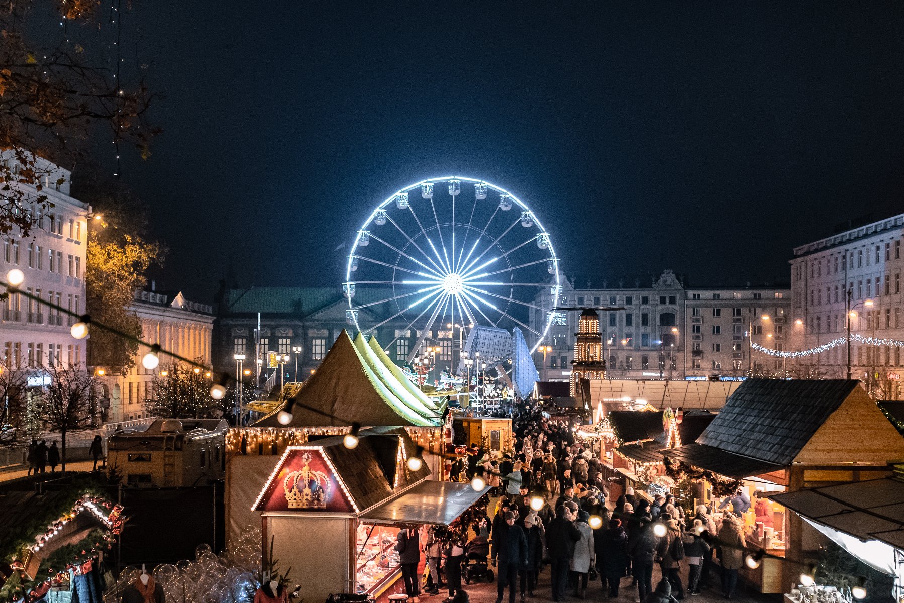 WIdok na Betlejem Poznańskie. Na pierwszym planie drewniane, rozświetlone budki. Między nimi spacerują ludzie ubrani w ciepłe kurtki, czapki i szaliki. W tle rozświetlony na biało Diabelski Młyn oraz stare kamienice. Niebo jest ciemne.