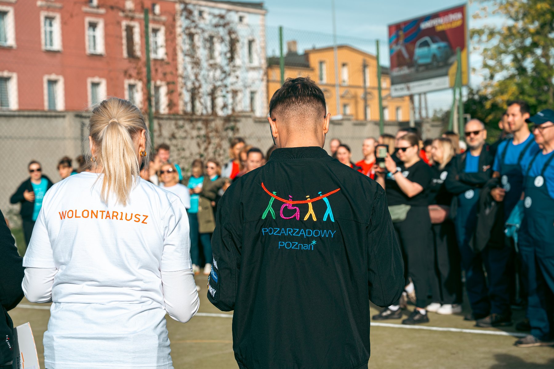 Widok na dwie osoby od tyłu, stojące na boisku, otoczone grupą ludzi. Kobieta po lewej ma na białej koszulce napis "WOLONTARIUSZ". Mężczyzna po prawej ma na czarnej kurtce kolorowe logo i napis w cudzysłowie: "POZARZĄDOWY POZNAŃ". W tle stare kamienice i duży billboard reklamowy.