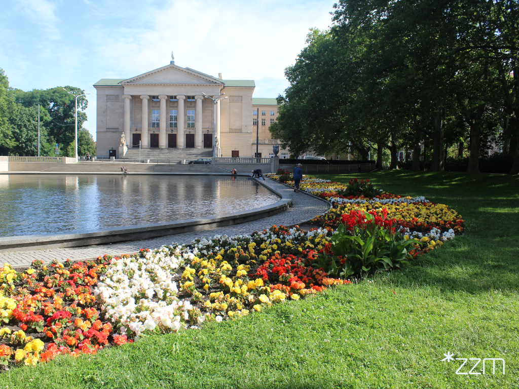 Widok na fontannę w parku Adama Mickiewicza i otaczające ją wielobarwne rabaty. W tle znajduje się gmach Teatru Wielkiego.