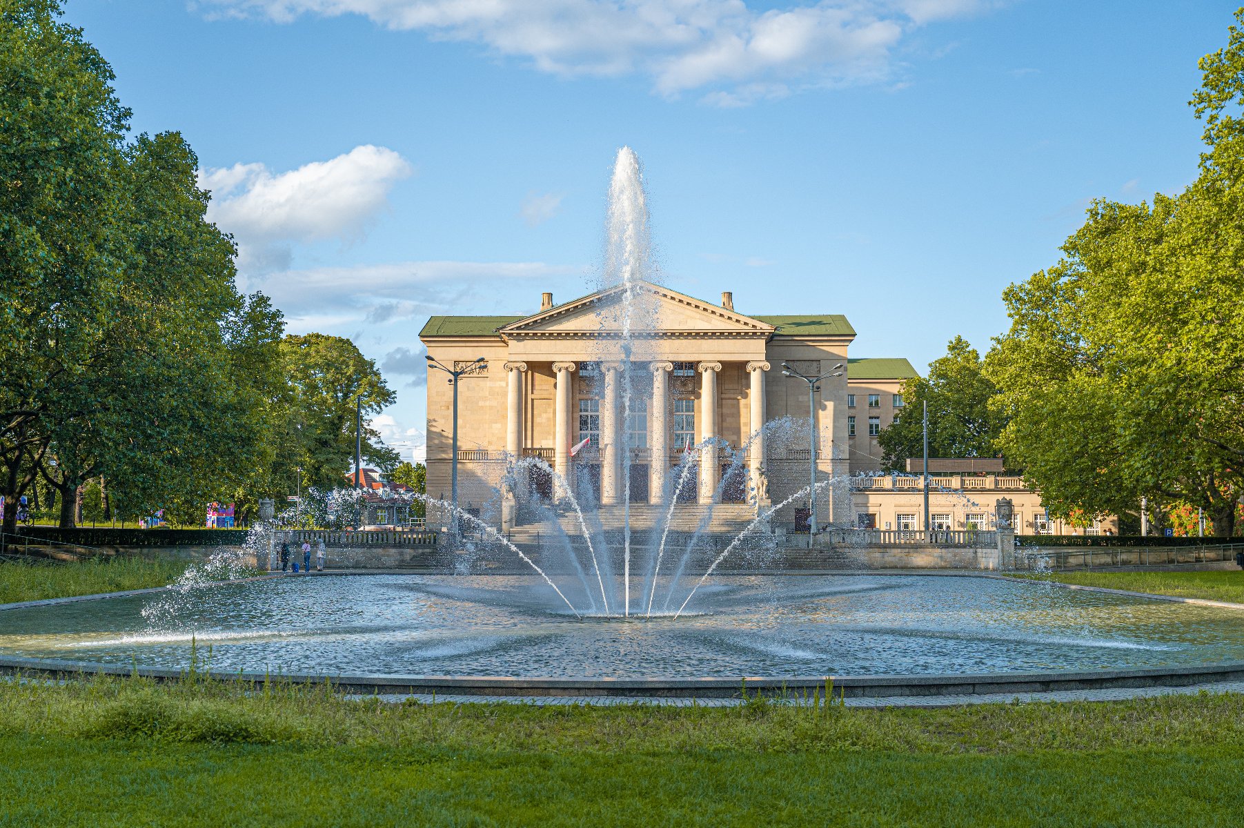 Widok na gmach Teatru Wielkiego im. Stanisława Moniuszki w Poznaniu, z fontanną na pierwszym planie. Fontanna wytryskuje wodę w kilku strumieniach, tworząc efektowną kurtynę wodną. Budynek teatru, utrzymany w klasycystycznym stylu, posiada kolumnowy portyk. Całość otoczona jest zielonym parkiem.