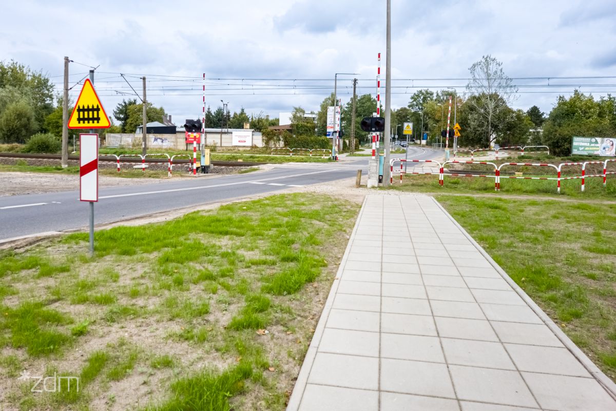 Widok na otwarty przejazd kolejowy, asfaltowa jezdnię, chodnik z płytek, biało-czerwone szlabany i barierki wzdłuż torów