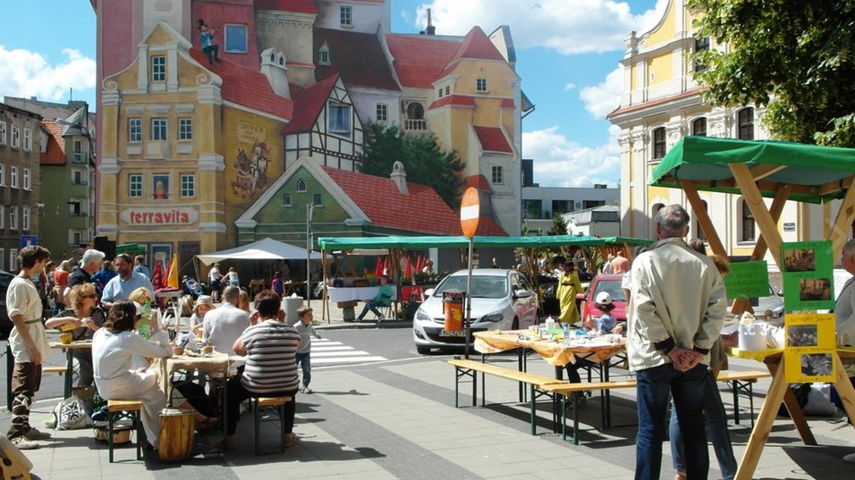 Widok na ryneczek na Śródce z rozłożonymi ławkami i stoiskami. W tle ściana ze słynym muralem