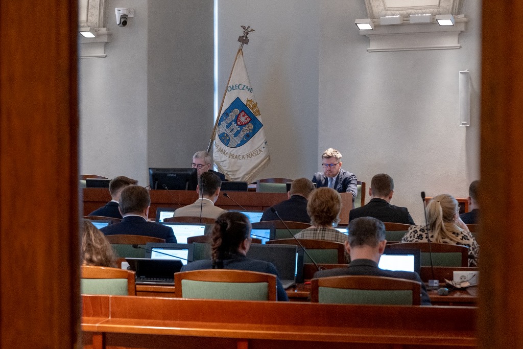 widok na salę sesyjna, przy stanowiskach radne i radni, przed nimi stół prezydialny, przy nim dwie osoby, za nimi sztandar z herbem Poznania