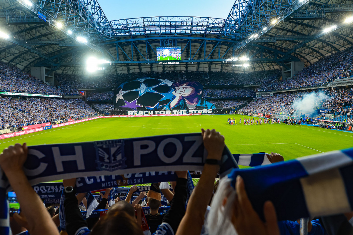 Widok na stadion Lecha Poznań wypełniony kibicami. Na pierwszym planie osoba trzymająca szalik Lecha. W tle sektorówka z napisem "reach For The Stars"