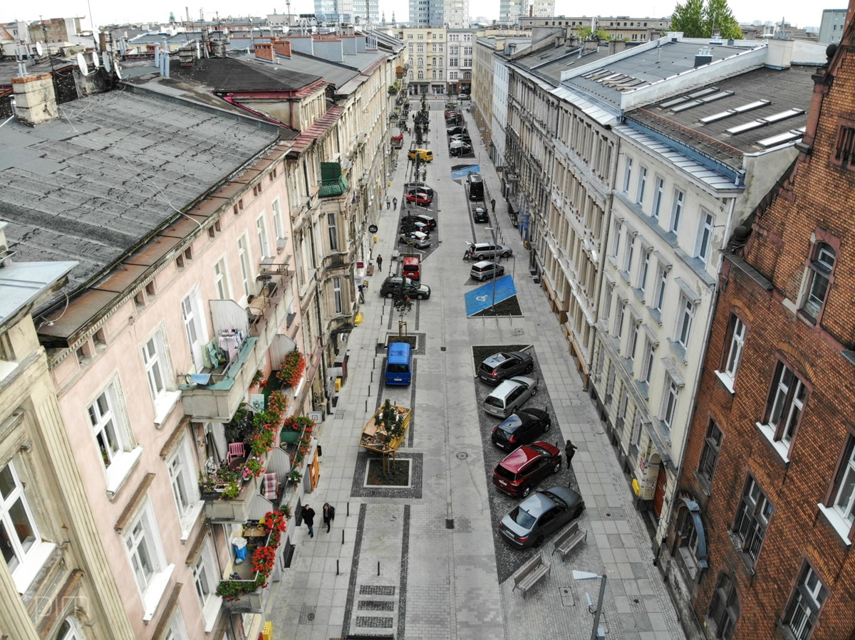 widok z dużej wysokości na wąską ulicę w zabudowie śródmiejskich kamienic, po prawej stronie samochody zaparkowane skośnie, po lewej kilka samochodów zaparkowanych równolegle do ulicy, pomiędzy ogródkami gastronomicznymi, są też miejsca parkingowe dla niepełnosprawnych