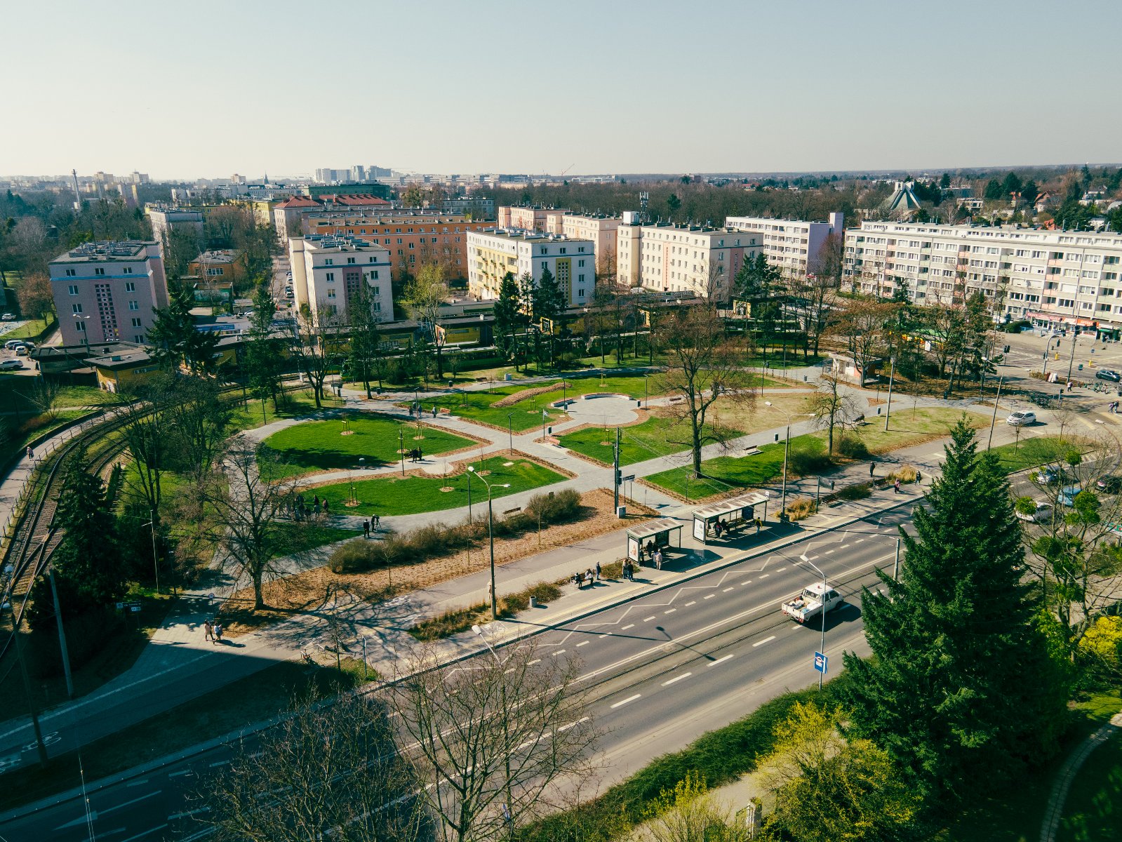 Widok z góry na Rondo Jeziorańskiego w Poznaniu. Pośrodku ronda znajduje się zielony park z alejkami i ławkami. Obok ronda biegnie ulica z ruchem samochodowym oraz tory tramwajowe. W tle widoczne są bloki mieszkalne i drzewa.