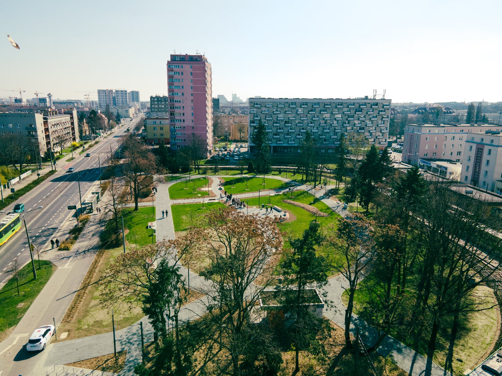 Widok z góry na skwer na Ogrodach. W centrum zdjęcia widać zielony park z alejkami i ławkami. Po lewej stronie parku biegnie ulica z ruchem samochodowym, a po prawej stronie widoczne są bloki mieszkalne i budynki.