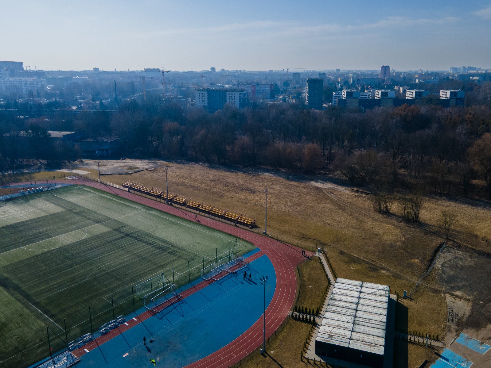Widok z lotu ptaka na boisko sportowe z bieżnią lekkoatletyczną. W tle drzewa oraz bloki