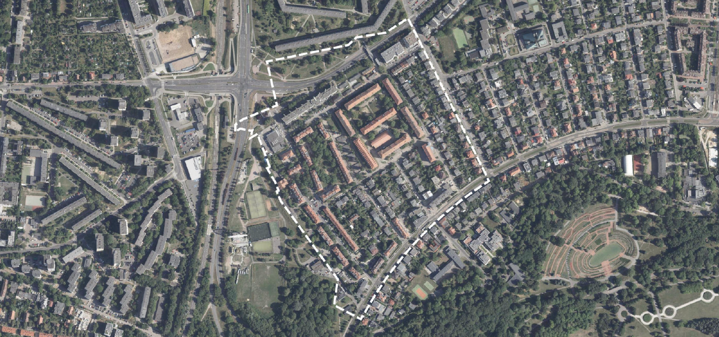 Widok z lotu ptaka na miejską przestrzeń. Obrazek ukazuje szczegółowy układ budynków, ulic i terenów zielonych. W centralnej górnej części krzyżuje się kilka głównych dróg, tworząc skomplikowaną siatkę ulic. W prawym dolnym rogu widoczny jest duży obszar zieleni z amfiteatrem lub okrągłą strukturą z tarasowymi siedzeniami. Zdjęcie oddaje zróżnicowanie funkcji miejskich - mieszkalnych, komercyjnych i rekreacyjnych.