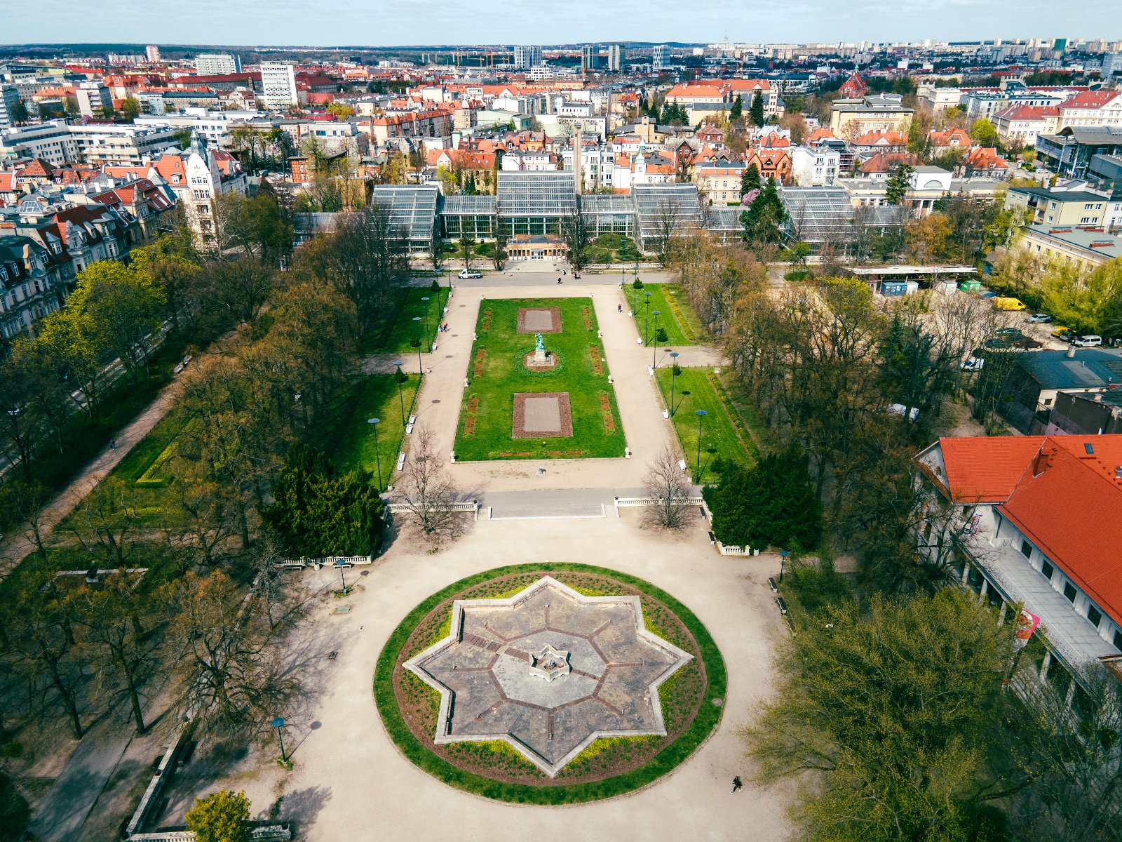 Widok z lotu ptaka na miejski park. Na pierwszym planie charakterystyczny, gwiaździsty element otoczony alejkami i zielenią. W centralnej części znajdują się prostokątne trawniki z pomnikiem pośrodku.