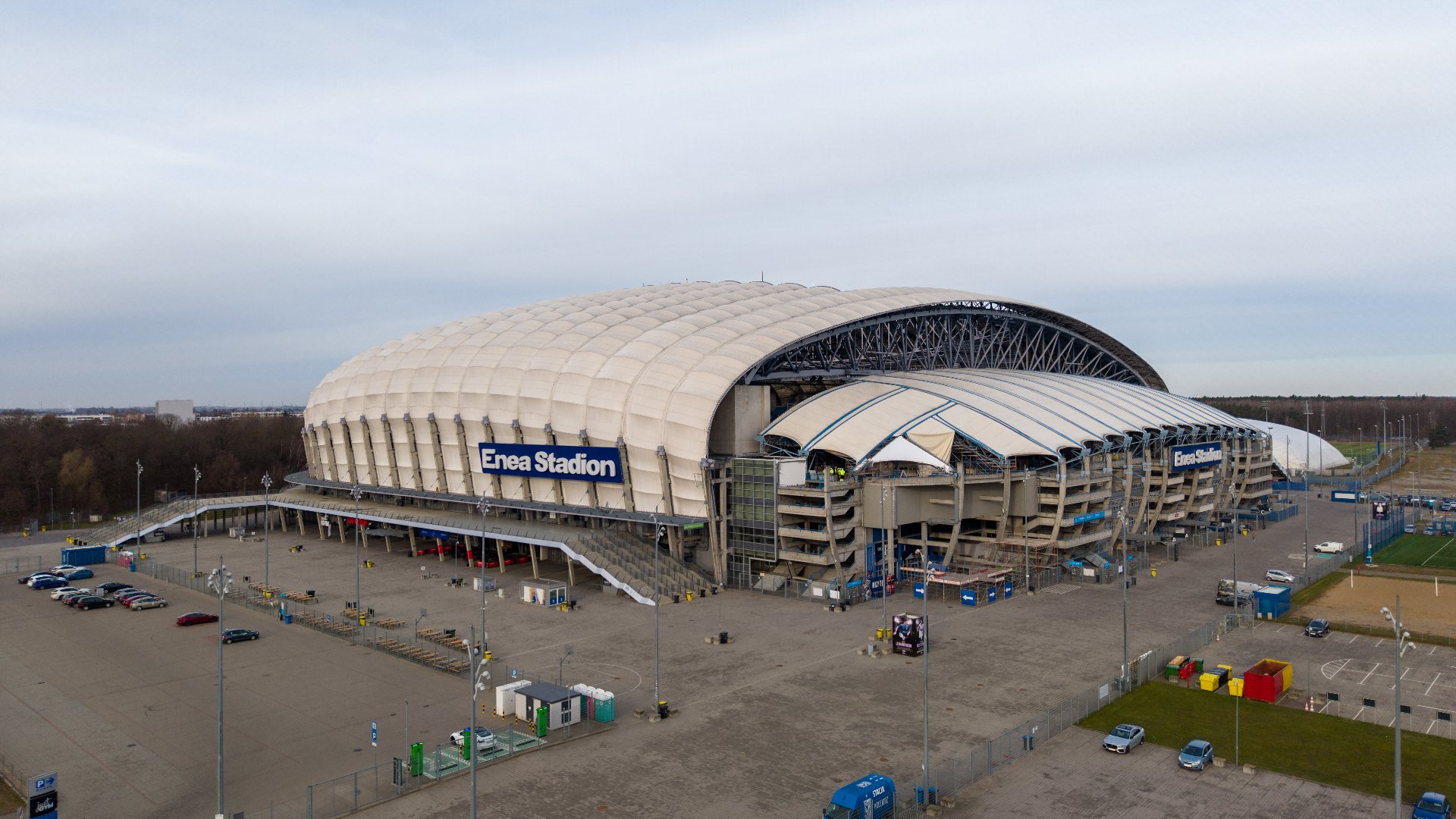Widok z lotu ptaka na obiekt Enea Stadion w Poznaniu wraz z otaczającymi go placami i parkingami pod lekko zachmurzonym niebem.