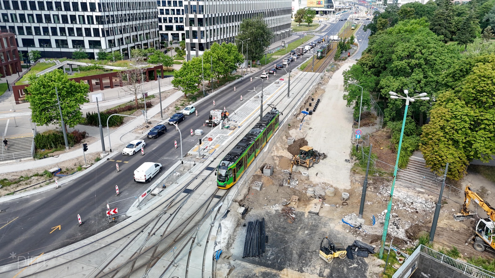 Widok z lotu ptaka na plac budowy. Pośrodku widać torowisko, a na nim jadący tramwaj. W tle samochody, wysokie budynki miejskie i zieleń parku.