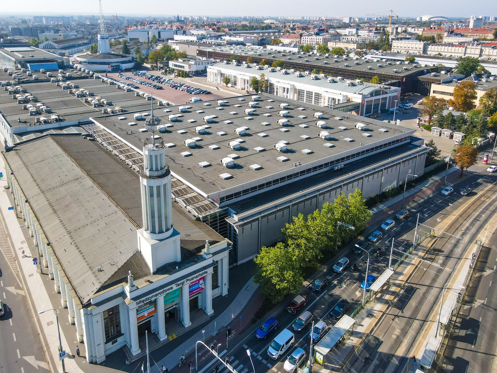 Widok z lotu ptaka na rozległy kompleks budynków, przypominający centrum wystawowe lub konferencyjne. Obiekty posiadają płaskie dachy z licznymi świetlikami i urządzeniami wentylacyjnymi. Jeden z budynków wyróżnia się klasyczną architekturą z kolumnami przy wejściu oraz wieżą ze spiczastą iglicą. W okolicy znajdują się parkingi, ulice z samochodami oraz inne budynki.