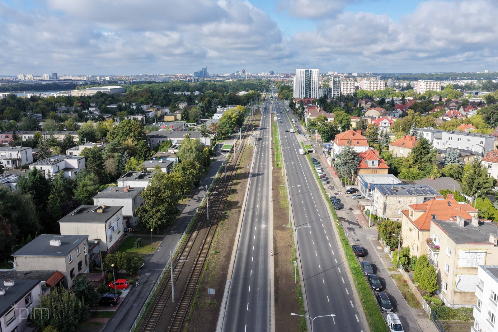Widok z lotu ptaka na szeroką miejską arterię z kilkoma pasami ruchu oraz torowiskiem tramwajowym biegnącym pośrodku jezdni. Po obu stronach ulicy znajdują się domy jednorodzinne, niewielkie budynki mieszkalne i liczne drzewa. W tle widać wysokie bloki oraz panoramę miasta. Niebo jest częściowo zachmurzone.