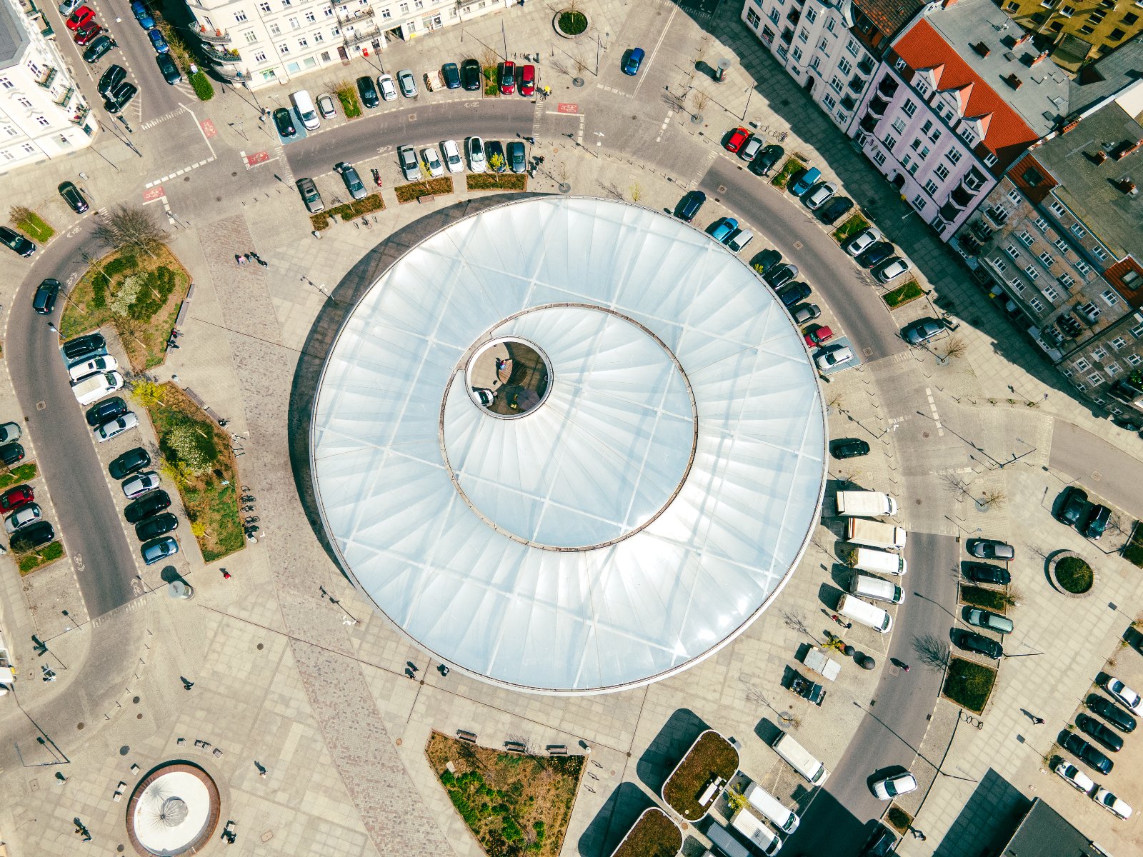 Widok z lotu ptaka. W centrum zdjęcia okrągła konstrukcja z przezroczystym dachem, otoczona placem i ulicami. Dookoła zaparkowane samochody i budynki mieszkalne. W dolnym lewym rogu fontanna i niewielki skwer.