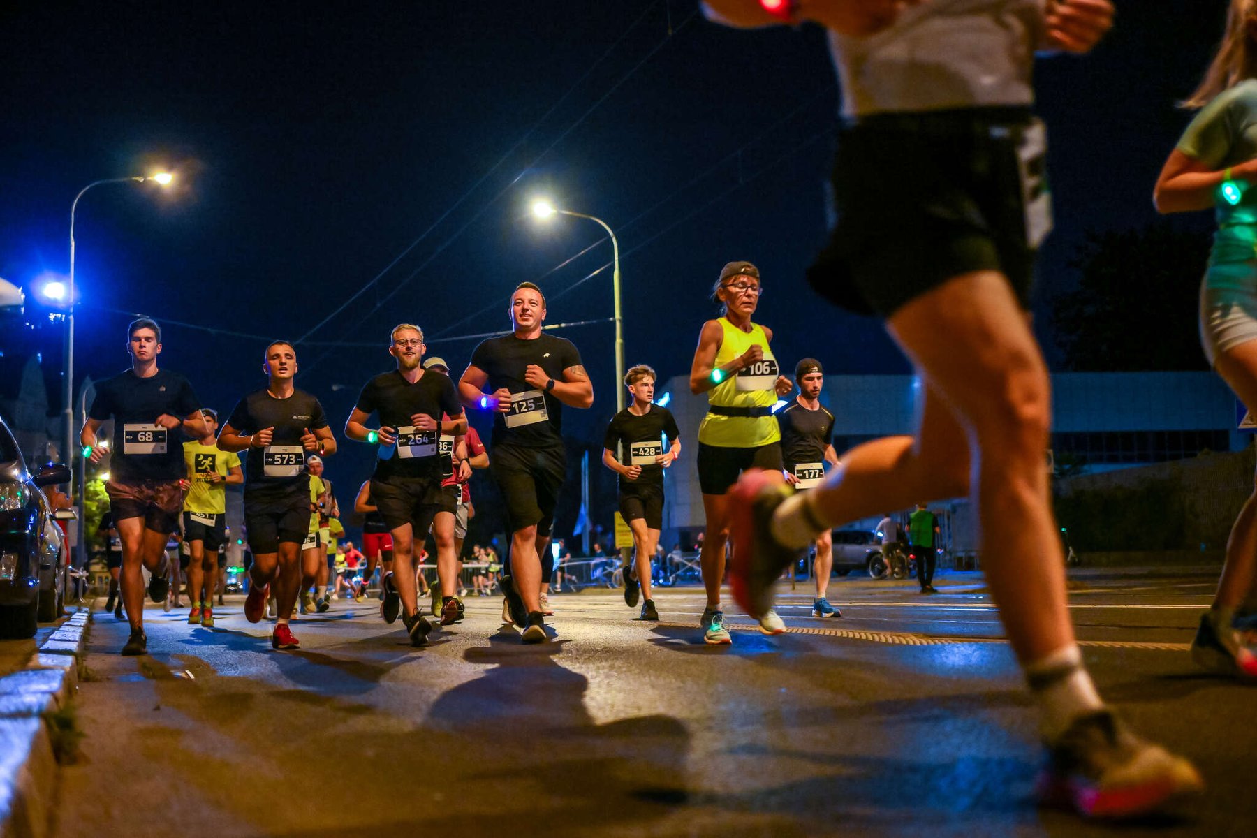 Wieczorny bieg uliczny, w którym biorą udział liczni zawodnicy. Wszyscy ubrani są w sportowe stroje, a niektórzy mają numery startowe. Mężczyzna w czarnej koszulce z numerem 1251 uśmiecha się. Po bokach drogi stoją latarnie uliczne, a w tle widać samochody.