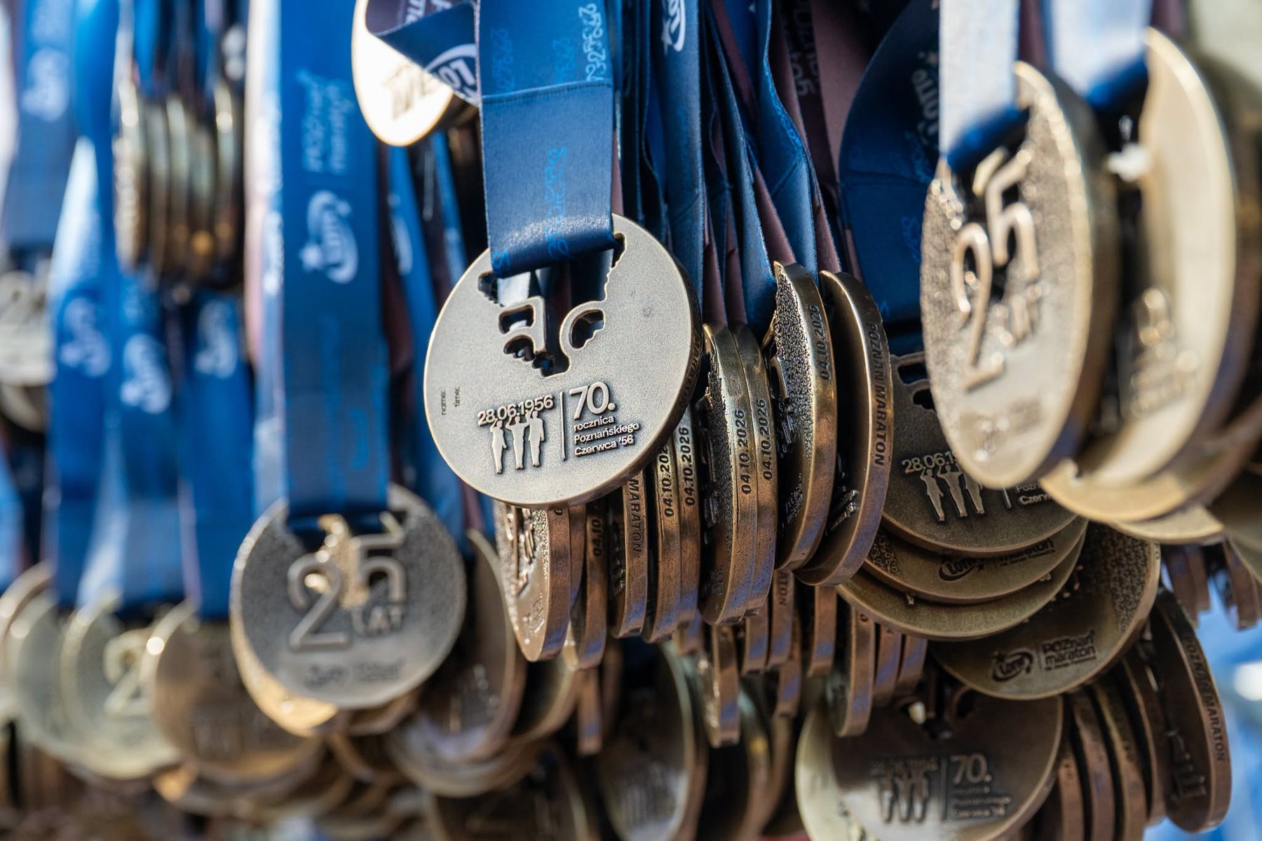 Wiele miedzianych metali wisi na niebieskich smyczach. Na awersie grawer "25 lat poznań maraton. Kamera skupia się na rewersie z symbolem rocznicy Czerwca 1956. Wygrawerowane 3 sylwetki niosące datę "28.06.1956".