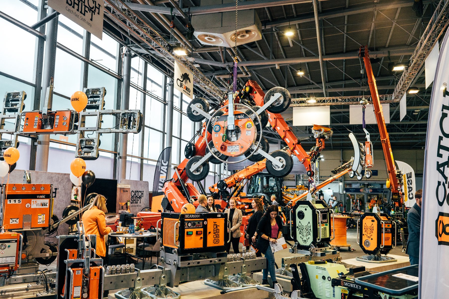 Wnętrze dużej hali targowej z ekspozycją ciężkich maszyn budowlanych i podnośników próżniowych marek VIAVAC oraz CATCH SHIFT. Na pierwszym planie pomarańczowe i zielone urządzenia z grafiką wilka, w głębi ramię dźwigu trzymające duży chwytak z przyssawkami. Wśród maszyn spacerują odwiedzający, widoczne są również pomarańczowe i czarne balony.