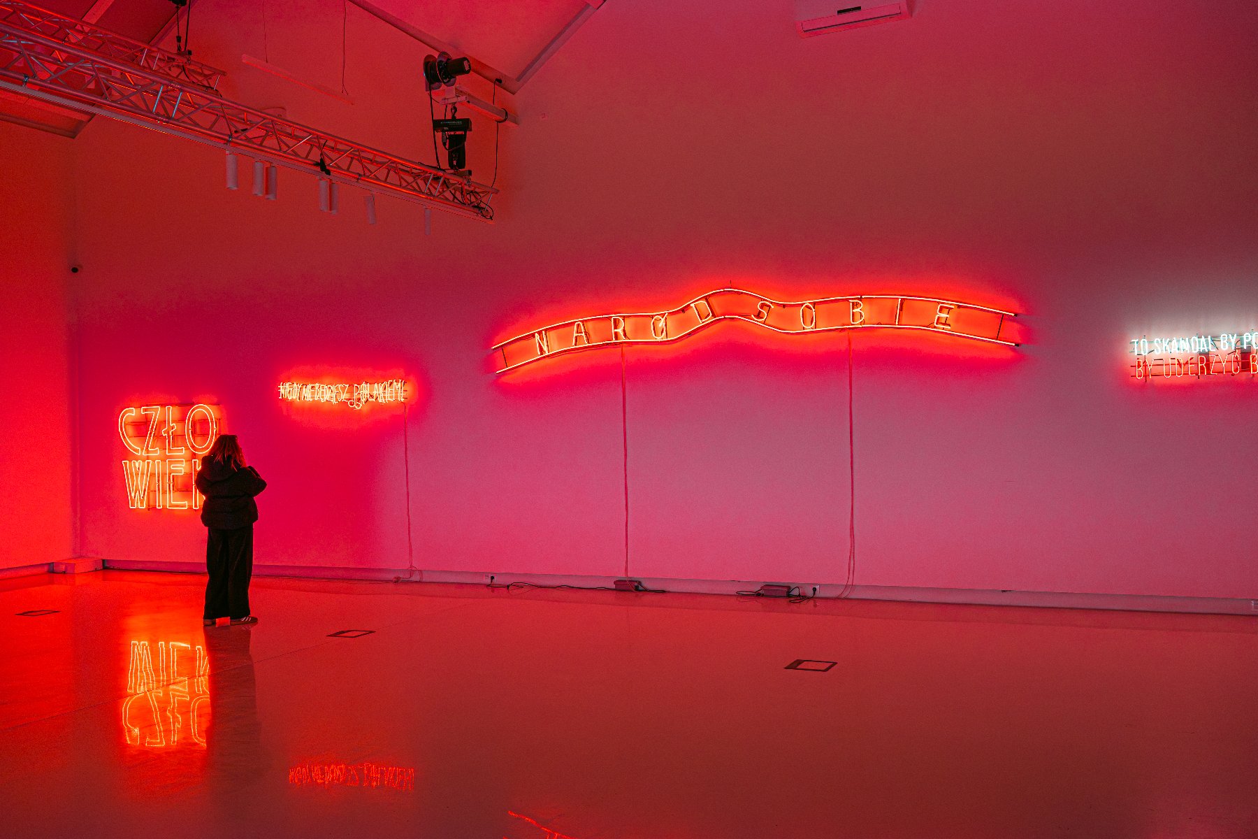 Wnętrze galerii sztuki skąpane w czerwonym świetle. Na białej ścianie zawieszone są cztery neonowe instalacje. Od lewej napisy: "CZŁOWIEK", mniejszy nieczytelny napis, duży falujący neon "NARÓD SOBIE" oraz tekst po prawej: "TO SKANDAL BY POSŁA UDERZYĆ B...". Przed neonami stoi tyłem postać w ciemnym ubraniu, której sylwetka odbija się w błyszczącej podłodze. Pod sufitem widoczna jest metalowa konstrukcja z oświetleniem.