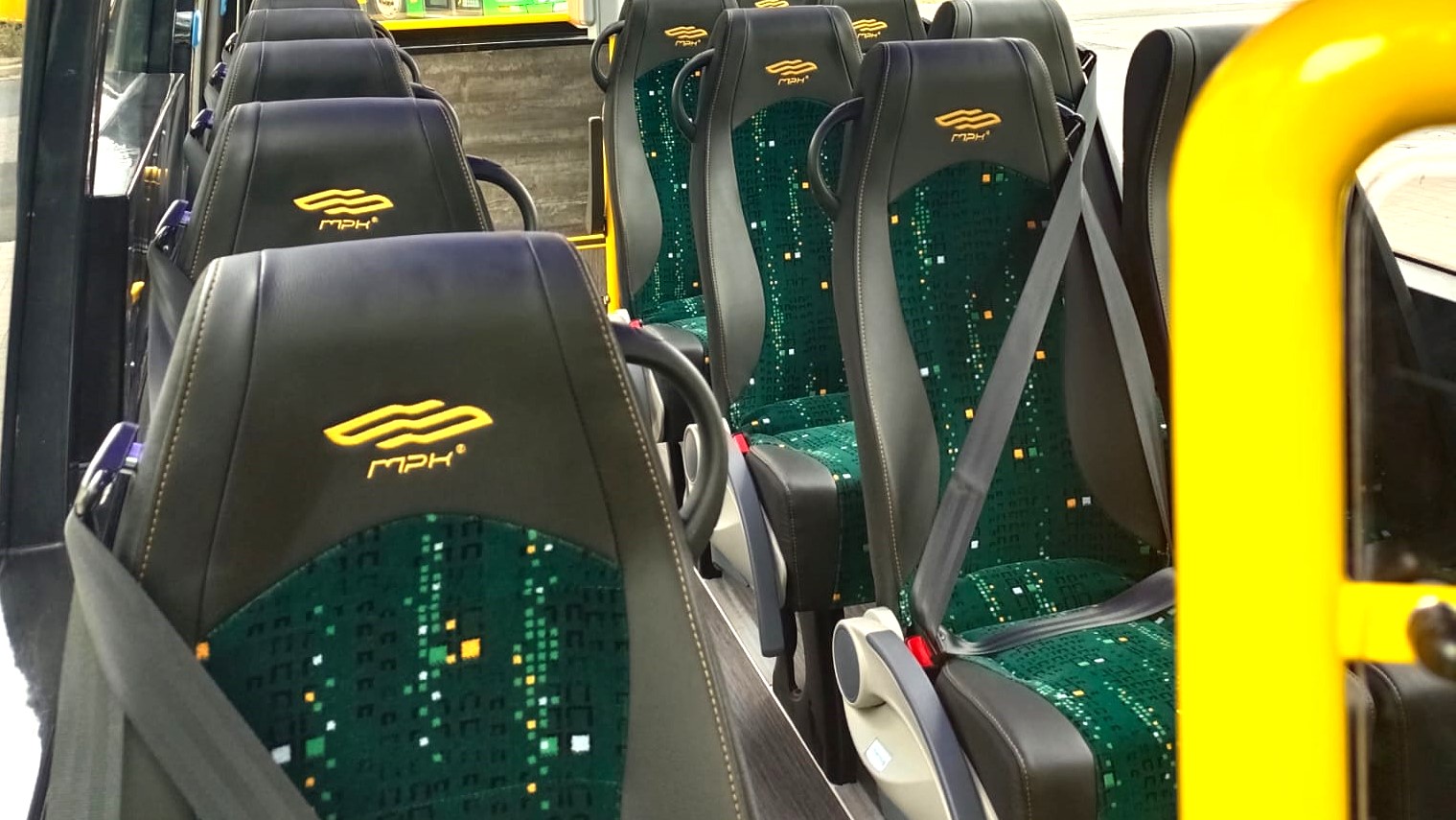 Wnętrze minibusa z rzędami siedzeń. Siedzenia są w czarno-zielone wzory, mają zagłówki i pasy bezpieczeństwa. Na zagłówkach widoczne jest logo MPK Poznań.