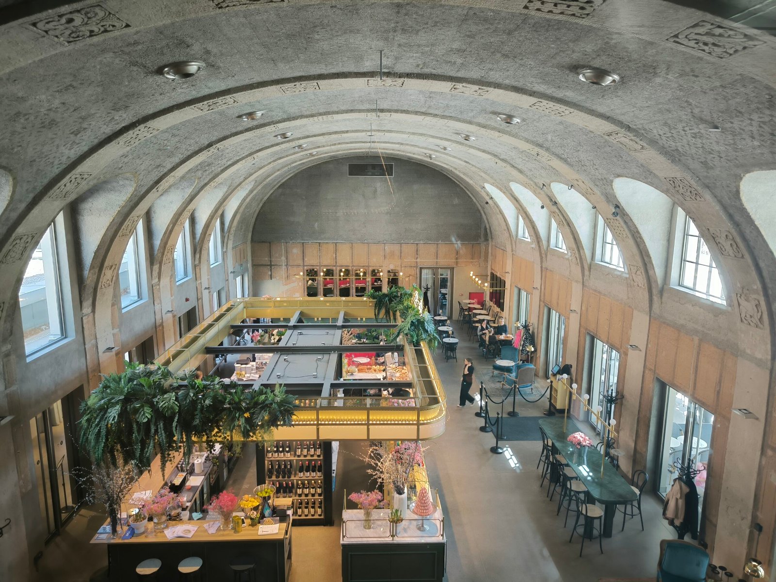 Wnętrze modernistycznego budynku z wysokim, łukowatym sufitem i rzędami okien, mieszczącego restaurację lub kawiarnię. W centralnej części widać otwartą kuchnię lub bar z wiszącymi roślinami i oświetleniem. Rozstawione są stoliki i krzesła, a w tle widać wejście i dalszą część lokalu. Architektura wnętrza sugeruje historyczny budynek, przekształcony na cele gastronomiczne.