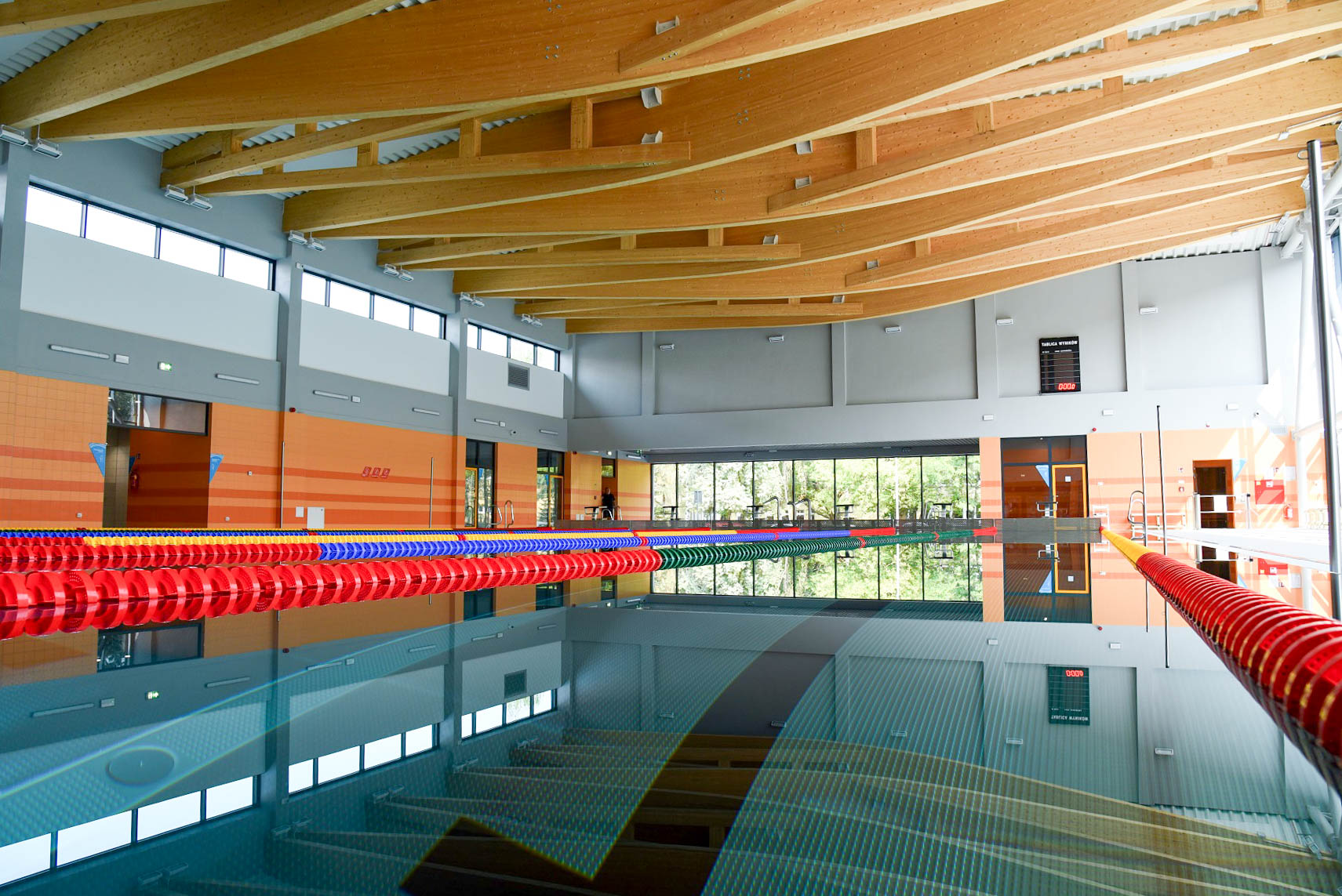 Wnętrze nowoczesnej pływalni z basenem sportowym, kolorowymi linami torowymi oraz efektownym zadaszeniem z drewnianych belkowych dźwigarów.