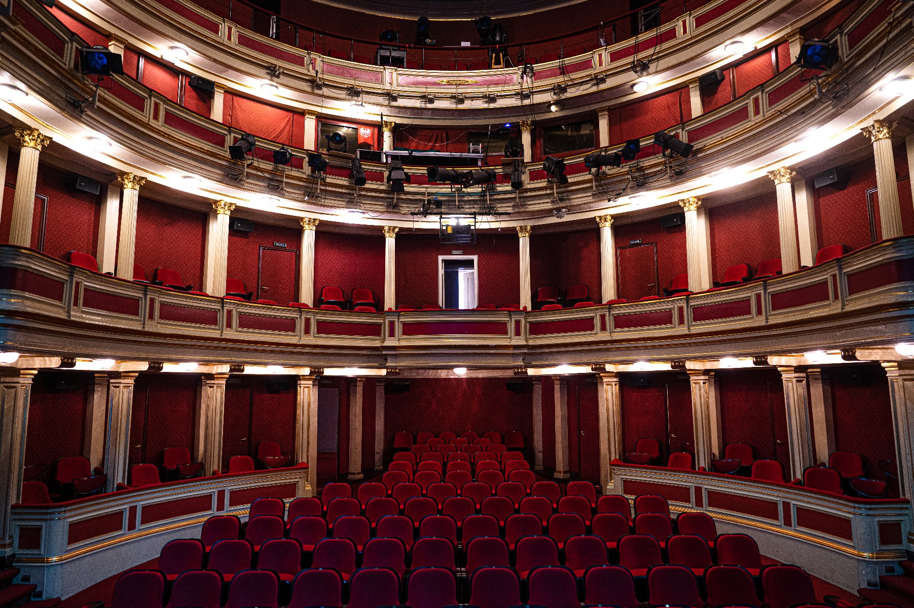 Wnętrze teatru. Widoczny parter oraz dwa balkony. Fotele z czerwonym obiciem, ściany zdobione czerwonymi panelami i złotymi akcentami. Balkony podtrzymywane przez ozdobne kolumny. Na balkonach zamontowane są różne urządzenia oświetleniowe i dźwiękowe. Teatr jest pusty, bez obecności widowni. Projekt elegancki i klasyczny, sugerujący tradycyjny lub historyczny teatr.