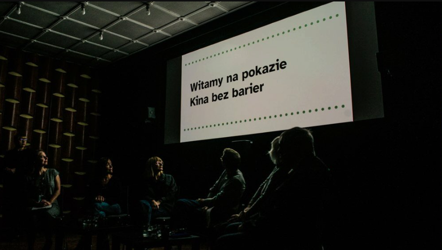 Wnętrze zaciemnionej sali kinowej. Na dużym ekranie widnieje czarny napis na białym tle: »Witamy na pokazie Kina bez barier«, ujęty w dwie poziome linie z zielonych kropek. Poniżej ekranu, w półmroku, siedzi grupa osób odwrócona tyłem lub bokiem do obiektywu, patrząca w stronę wyświetlanego komunikatu.