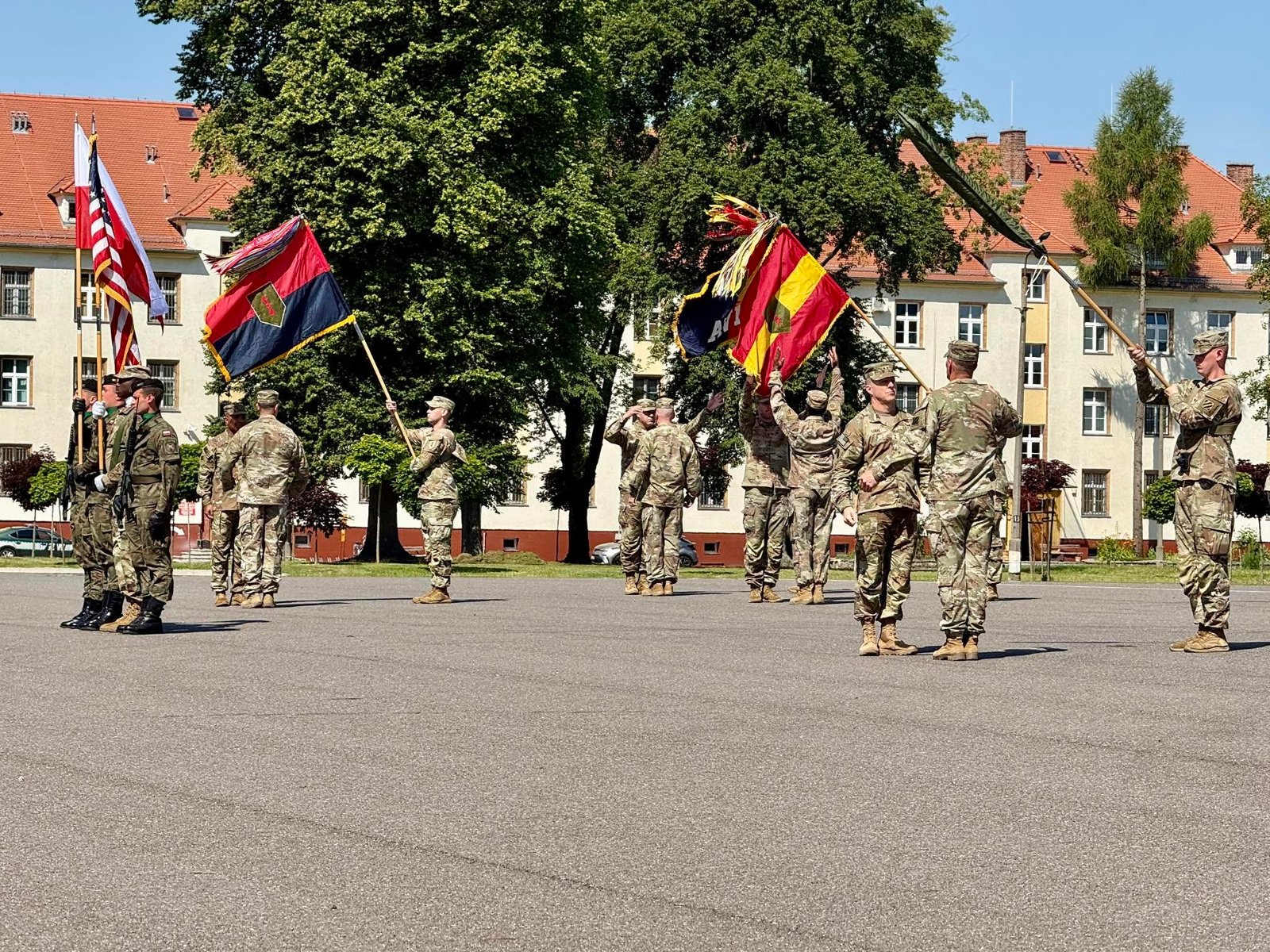 Wojskowa uroczystość na placu apelowym z udziałem amerykańskich i polskich żołnierzy. Widoczne są poczty sztandarowe z flagami narodowymi (Polski i USA) oraz proporcami jednostek wojskowych, a w tle stoją budynki koszarowe otoczone drzewami.