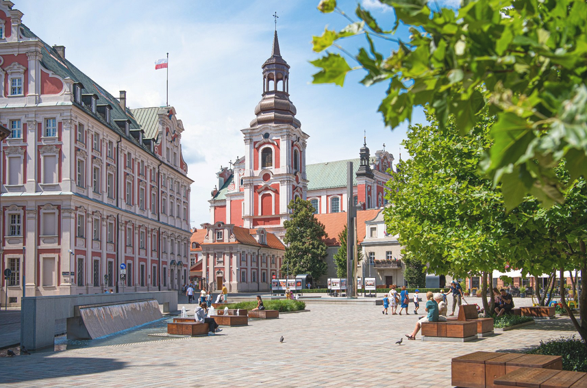wybrukowany skwer, ludzie siedzą na ławkach lub spacerują, w tle wzniosłe gmachy, na jednym z nich powiewa biało-czerwona flaga