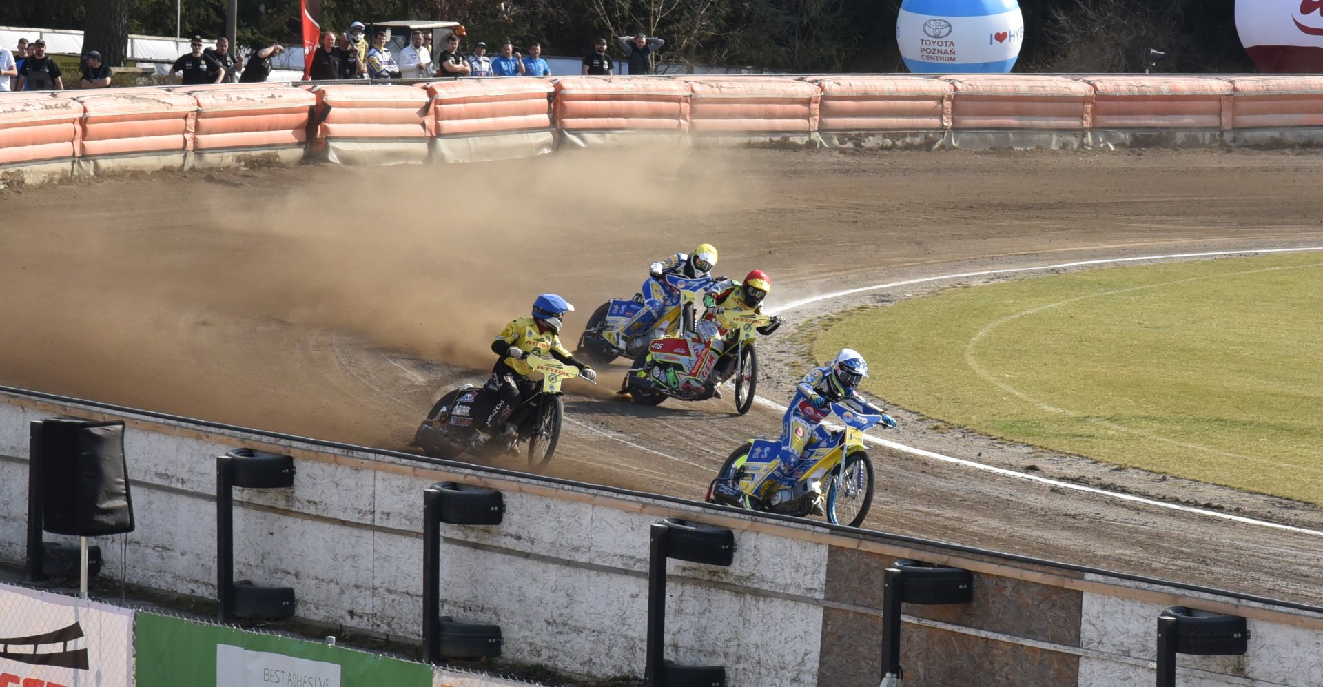 Wyścig motocyklowy na torze ziemnym. Na pierwszym planie cztery motocykle z zawodnikami w kaskach i strojach ochronnych. Tor otoczony barierkami, w tle widoczni są widzowie obserwujący zmagania. Z motocykli unosi się kurz, który nadaje dynamizmu całej scenie. Na barierkach reklamowych znajdują się różne banery, w tym z logo Toyota. Widoczne emocje i rywalizacja podkreślają napięcie wyścigu.