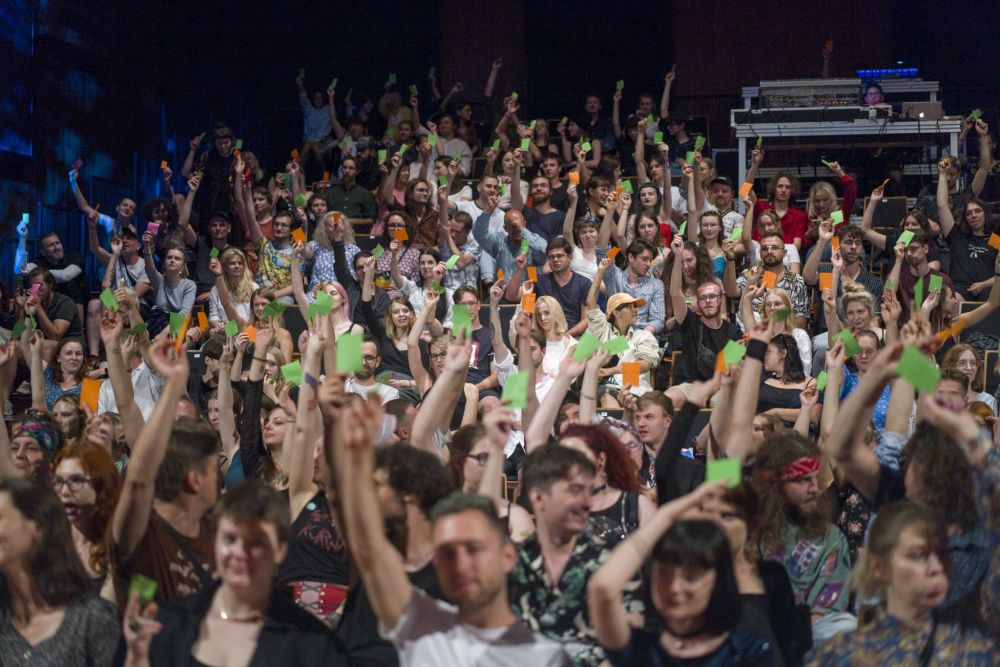 Występująca oświetlona scenicznym światłem jest odwórcona do publizności, która głosuje, pokazując niebieskie lub fioletowe karteczki.
