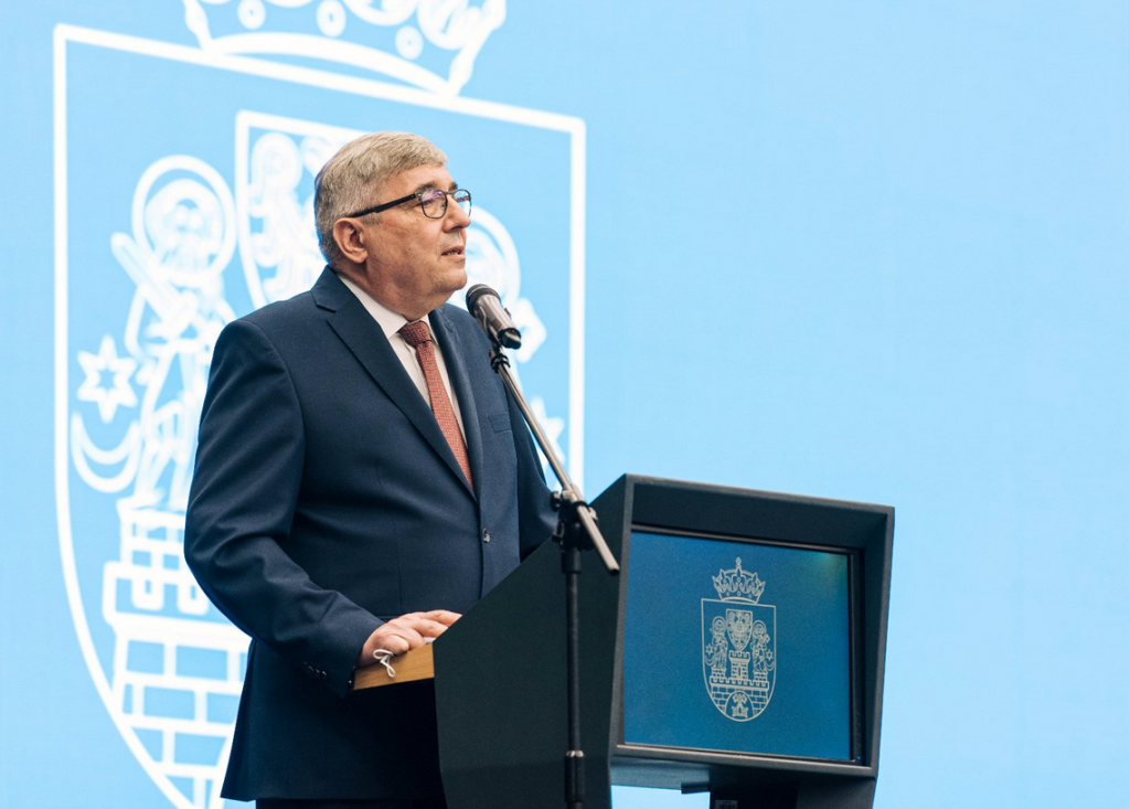 za mówniącą z mikrofonem stoi mężczyzna w garniturze, krawat i jasna koszula, okulary, włosy krótkie zaczesane na bok, częściowo siwiejące, z przodu mównicy herb Miasta Poznania, za mężczyzną ściana z dużym herbem Poznania