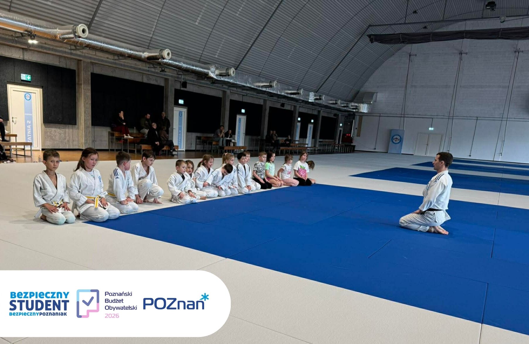 Zajęcia w ramach projektu "Bezpieczny Student - Bezpieczny Poznaniak" - trener siedzi na macie, a zawodnicy przed nim siedzą przed matą w hali