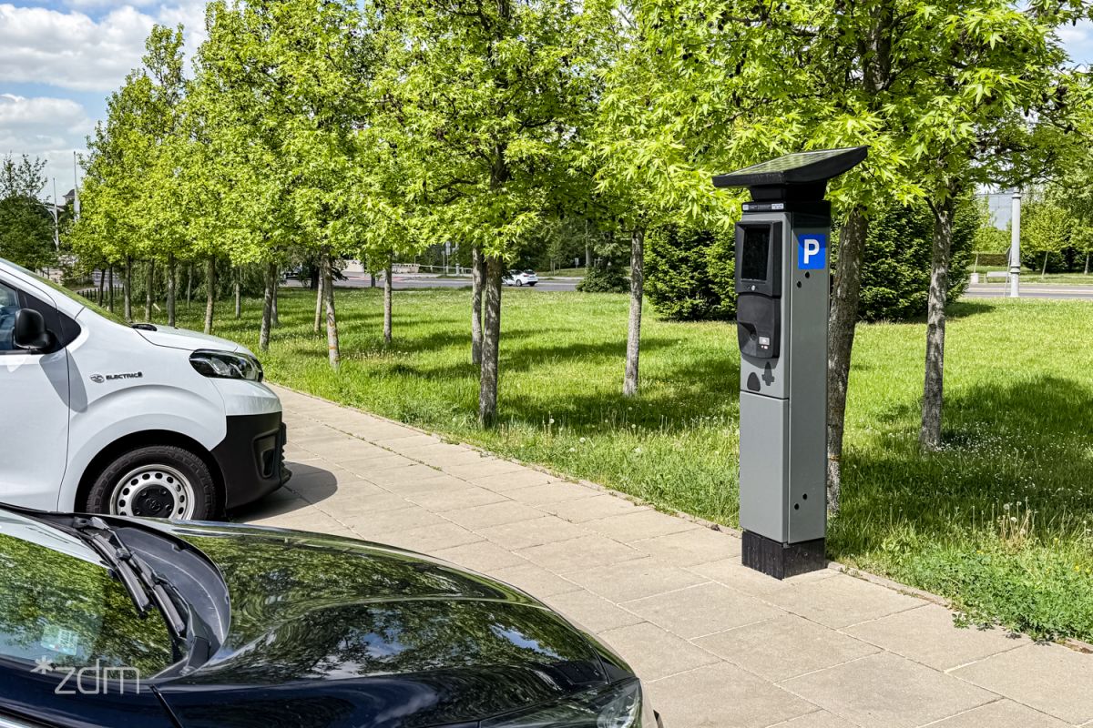 Zaparkowane samochody przy zielonym skwerze. Na chodniku stoi parkomat