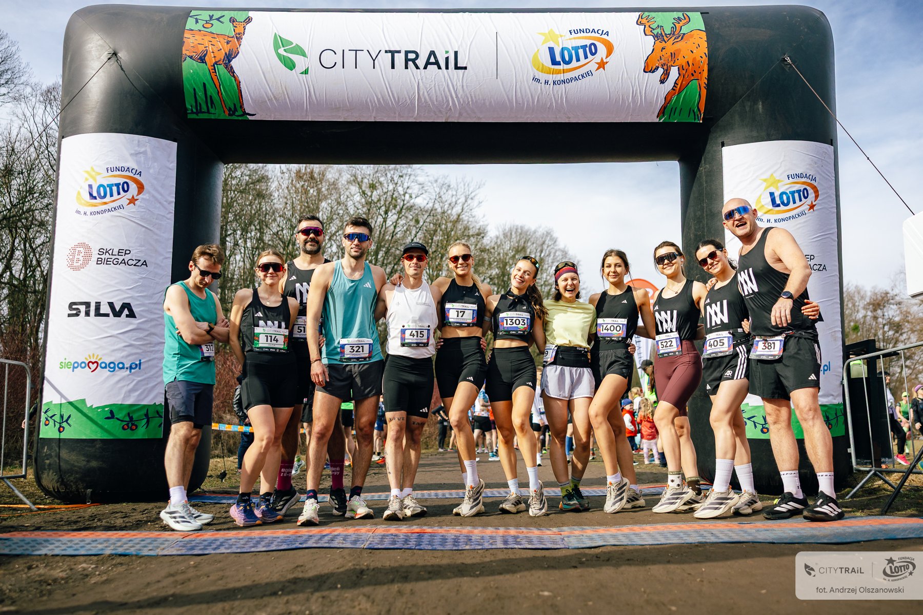 Zawodnicy przed startem w ostatnim biegu sezonu City Trail Poznań, fot. Andrzej Olszanowski - kilkanaścioro zawodników stoi przed bramą startową zawodów. Uczestnicy są w letnich strojach, przede wszystkim w czarnej odzieży. W tle widać las