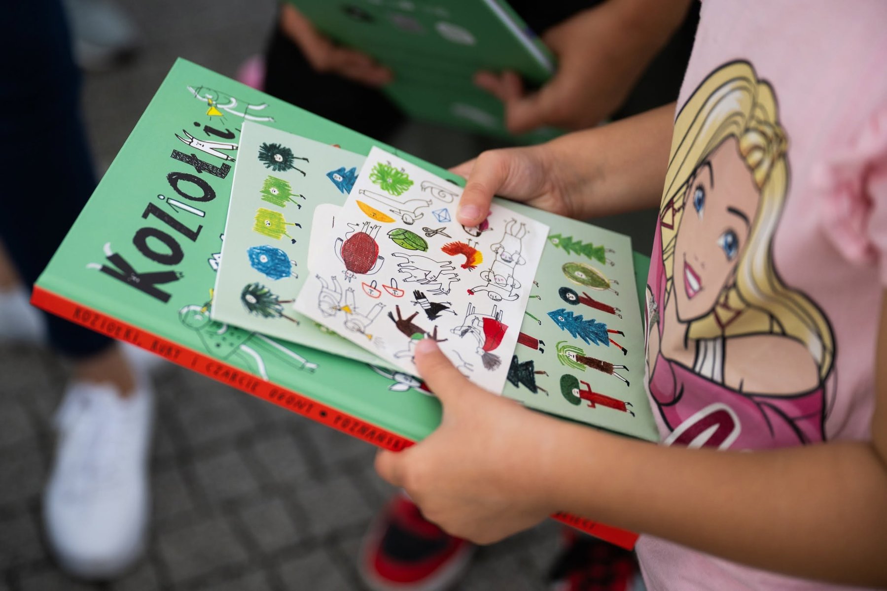 Zbliżenie na dłonie dziecka trzymającego zieloną książkę pod tytułem "Koziołki" oraz kolorowe pocztówki i naklejki z ilustracjami.