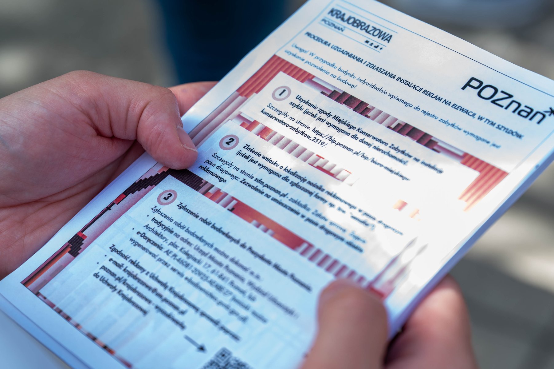 Zbliżenie na dłonie trzymające białą ulotkę informacyjną z logo "POZnan*" i nagłówkiem "KRAJOBRAZOWA POZNAŃ". Treść ulotki zawiera ponumerowane w kółkach punkty 1, 2 i 3, opisujące procedurę uzgadniania i zgłaszania instalacji reklam na elewacji. W tekście widoczne są m.in. słowa: "Uzyskanie zgody Miejskiego Konserwatora Zabytków", "Złożenie wniosku o lokalizację nośnika reklamowego" oraz "Zgłoszenie robót budowlanych".