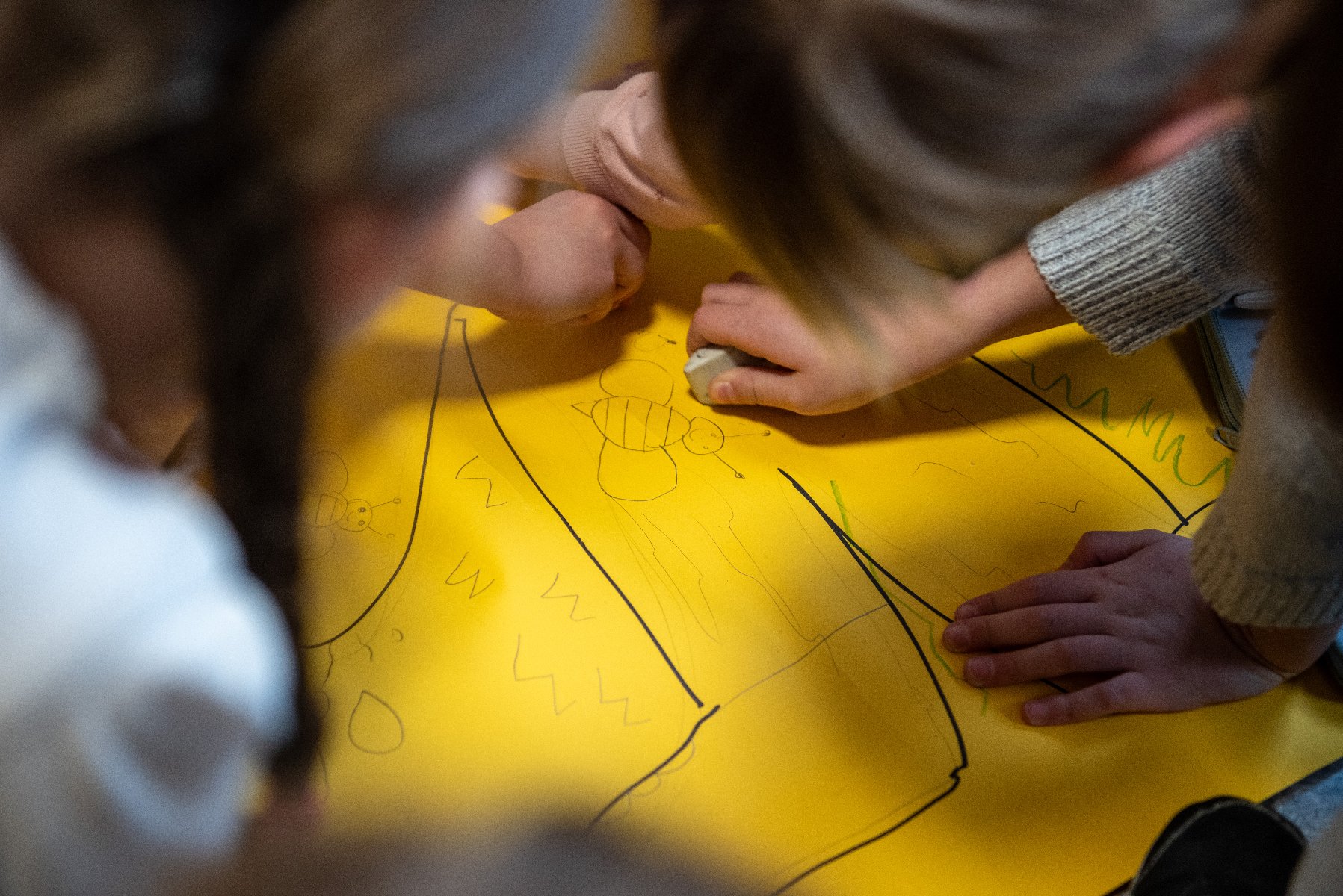 Zbliżenie na ręce dzieci pracujących wspólnie nad rysunkiem na dużym żółtym arkuszu papieru. Na papierze widoczne są kontury narysowanych ołówkiem pszczół oraz faliste linie. Jedno z dzieci używa gumki do mazania, aby poprawić fragment szkicu. Kadrujące ujęcie z góry podkreśla skupienie i wspólną aktywność twórczą.