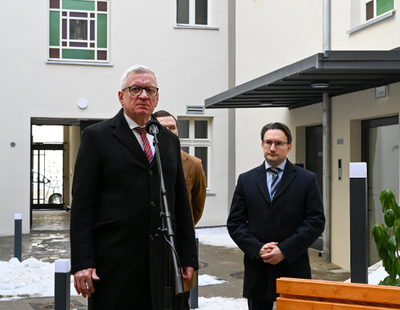 Zbliżenie na starszego mężczyznę w okularach i czarnym płaszczu z czerwonym krawatem, który przemawia do mikrofonu na statywie. Za nim, po prawej stronie, stoi nieco niższy mężczyzna w okularach i ciemnym płaszczu z rękami spllecionymi przed sobą.