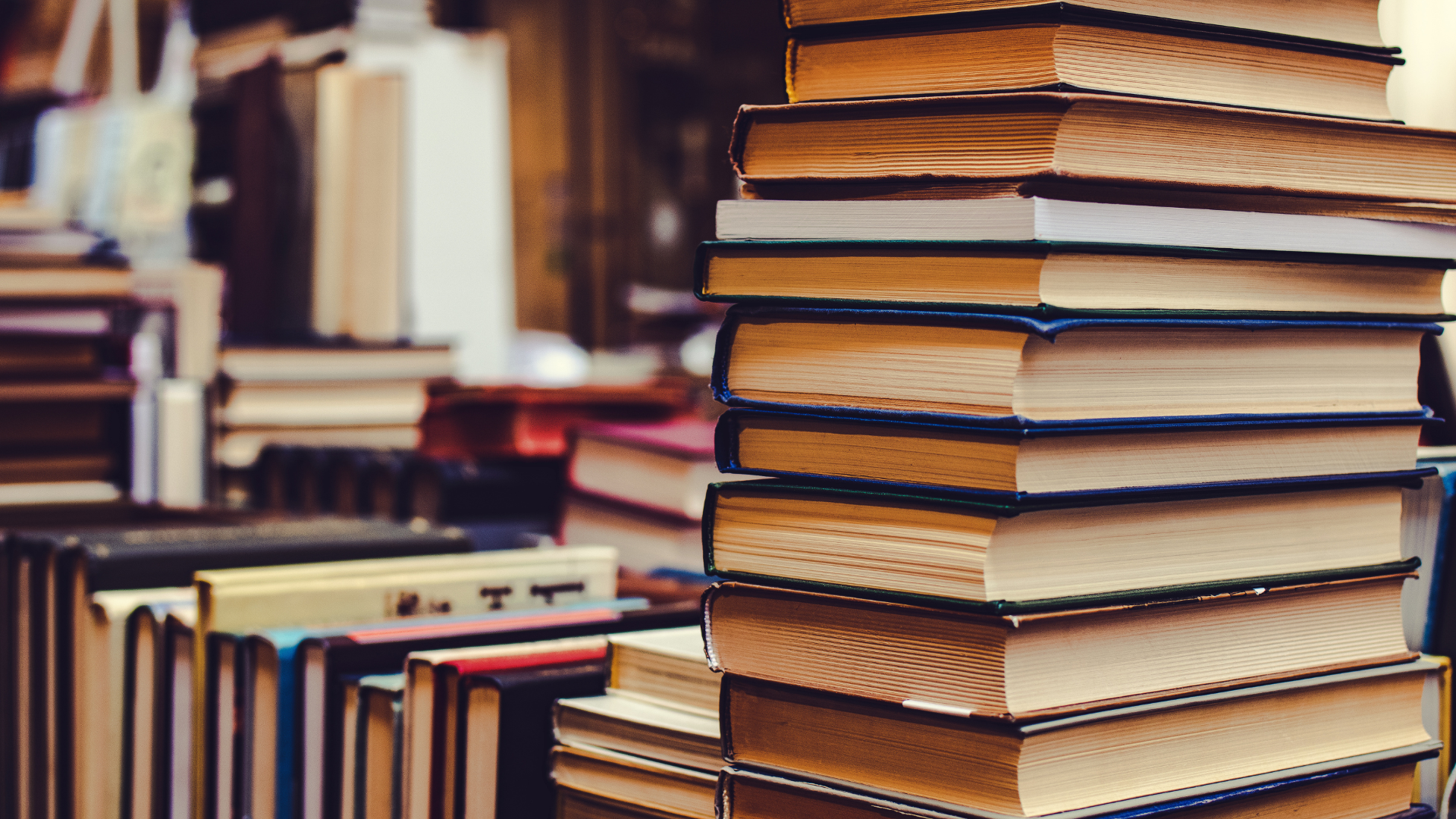 Zbliżenie na stos książek po prawej stronie kadru, z książkami ułożonymi w rzędy w tle. Dominują odcienie brązu i bieli.