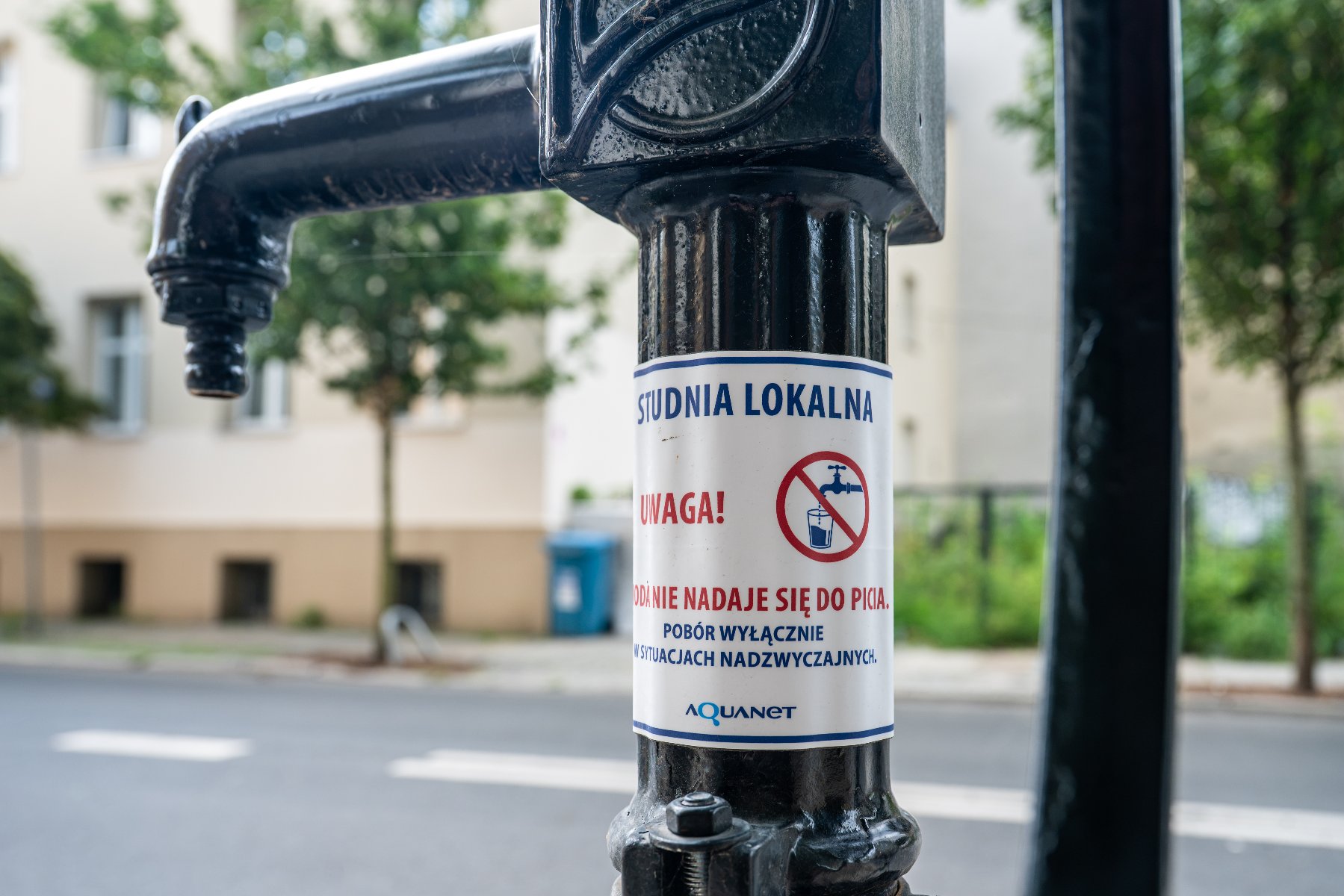 Zbliżenie na uliczną pompę wodną Aquanet z tabliczką ostrzegawczą informującą o studni lokalnej i braku zdatności wody do picia.