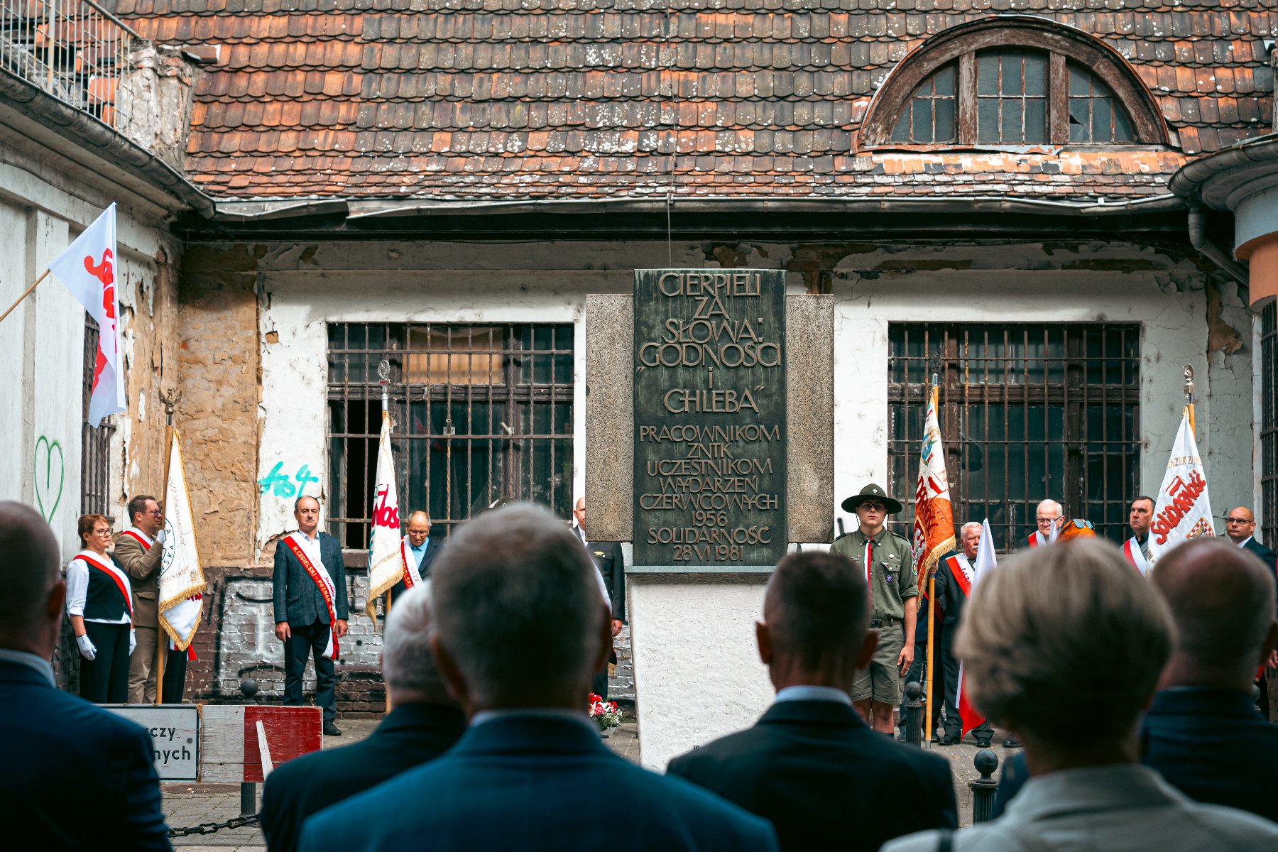 Zdjęcie dokumentuje uroczystość patriotyczną odbywającą się przed tablicą pamiątkową poświęconą uczestnikom Czerwca '56. Tablica z napisem "CIERPIELI ZA SŁOWO, GODNOŚĆ I CHLEBA" jest umieszczona na zniszczonej ścianie budynku z kratami w oknach. W ceremonii biorą udział poczty sztandarowe, harcerz oraz osoby w strojach oficjalnych z biało-czerwonymi szarfami.