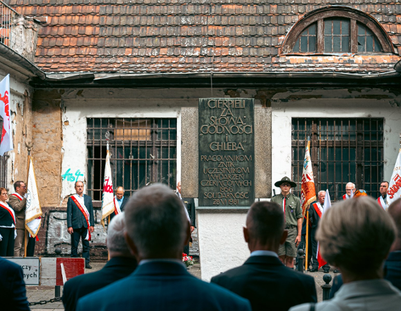 Zdjęcie dokumentuje uroczystość patriotyczną odbywającą się przed tablicą pamiątkową poświęconą uczestnikom Czerwca '56. Tablica z napisem "CIERPIELI ZA SŁOWO, GODNOŚĆ I CHLEBA" jest umieszczona na zniszczonej ścianie budynku z kratami w oknach. W ceremonii biorą udział poczty sztandarowe, harcerz oraz osoby w strojach oficjalnych z biało-czerwonymi szarfami.