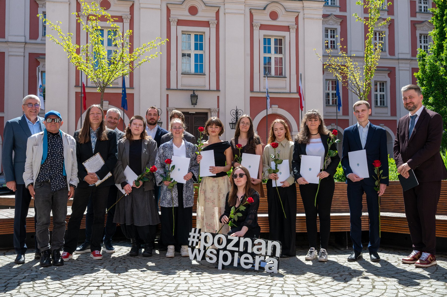Zdjęcie grupowe. Grupa kobiet i mężczyzn, tegorocznych laureatek i laureatów pozuje wspólnie z Kapitułą oraz wręczającymi stypendia. W tle nasłoneczniony dziedziniec Placu Kolegiackiego.