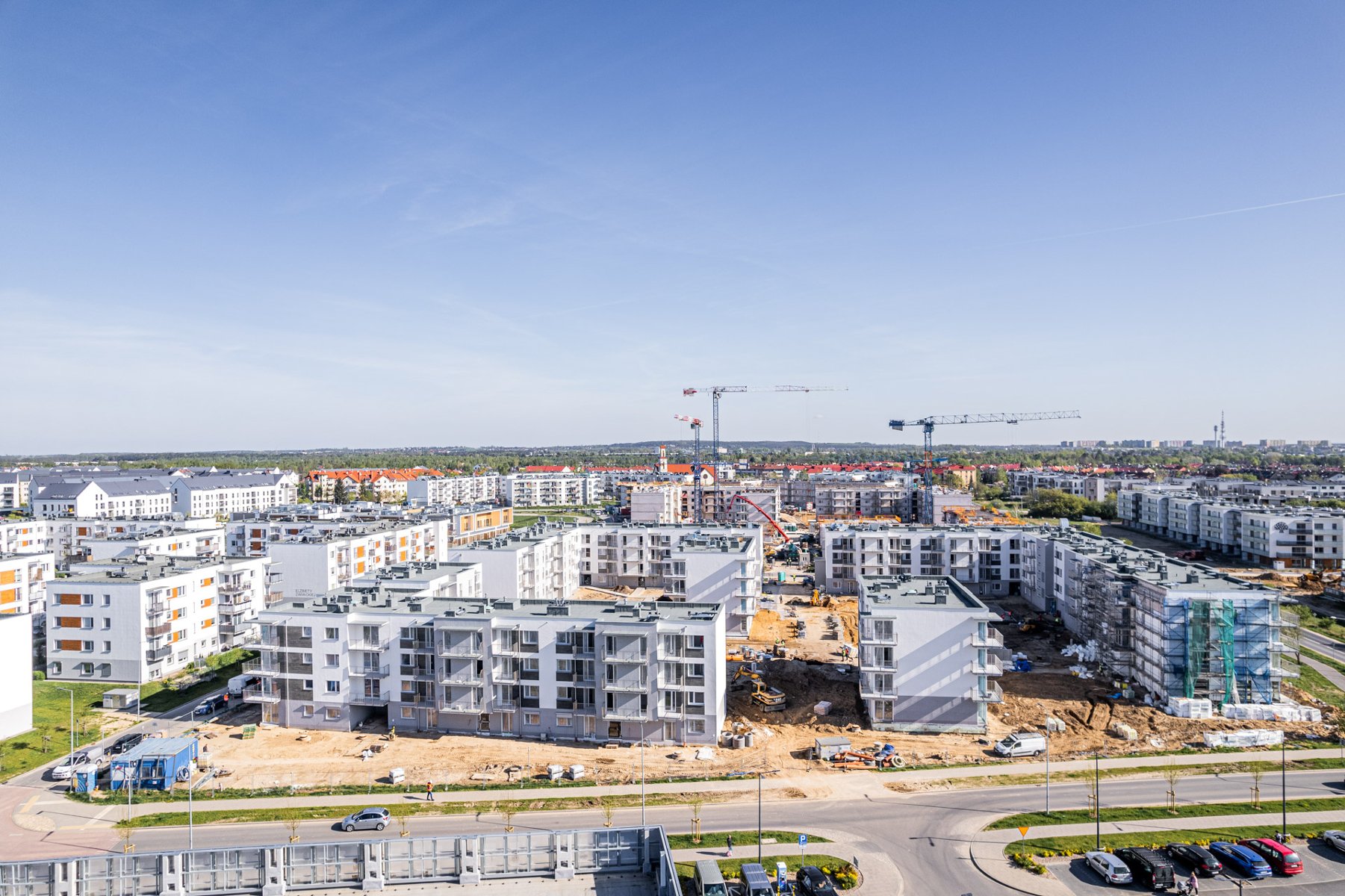 Zdjęcie lotnicze przedstawiające duży plac budowy osiedla mieszkaniowego, na którym wznoszone są kilkupiętrowe, jasne bloki. W centralnej części widoczne są pracujące żurawie budowlane oraz maszyny ziemne, a wokół znajdują się gotowe już budynki mieszkalne oraz drogi dojazdowe z parkingami.