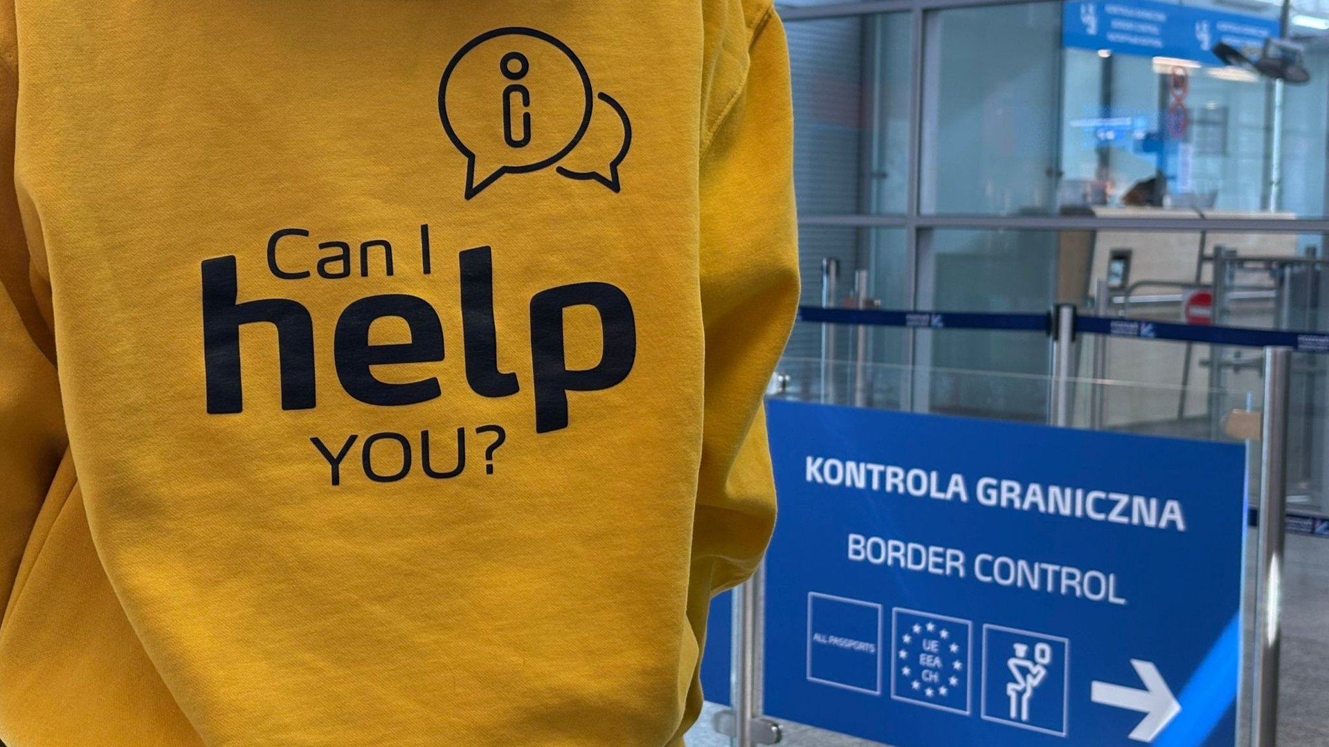 Zdjęcie osoby w żółtej bluzce z dużym czarnym napisem "Can I help you?" oraz ikoną dymku z symbolem informacji. W tle widoczna jest niebieska tablica na lotnisku z napisem "Kontrola graniczna" oraz strzałkami kierującymi podróżnych.